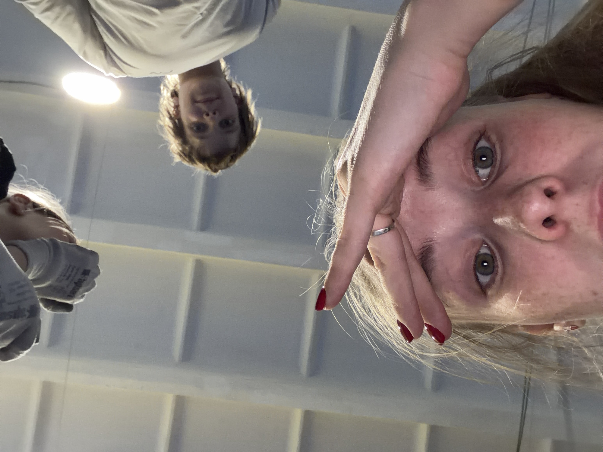 Three people are visible in the image. One person, wearing work gloves, is partially visible on the left side. A second person with short brown hair is visible near the center of the image. A third person, with long blonde hair and red nail polish, is at the right of the image. The location appears to be an indoor space with a white ceiling and a light fixture visible in the background. It is not possible to determine if the location is in Berezniki, Russia without additional information.