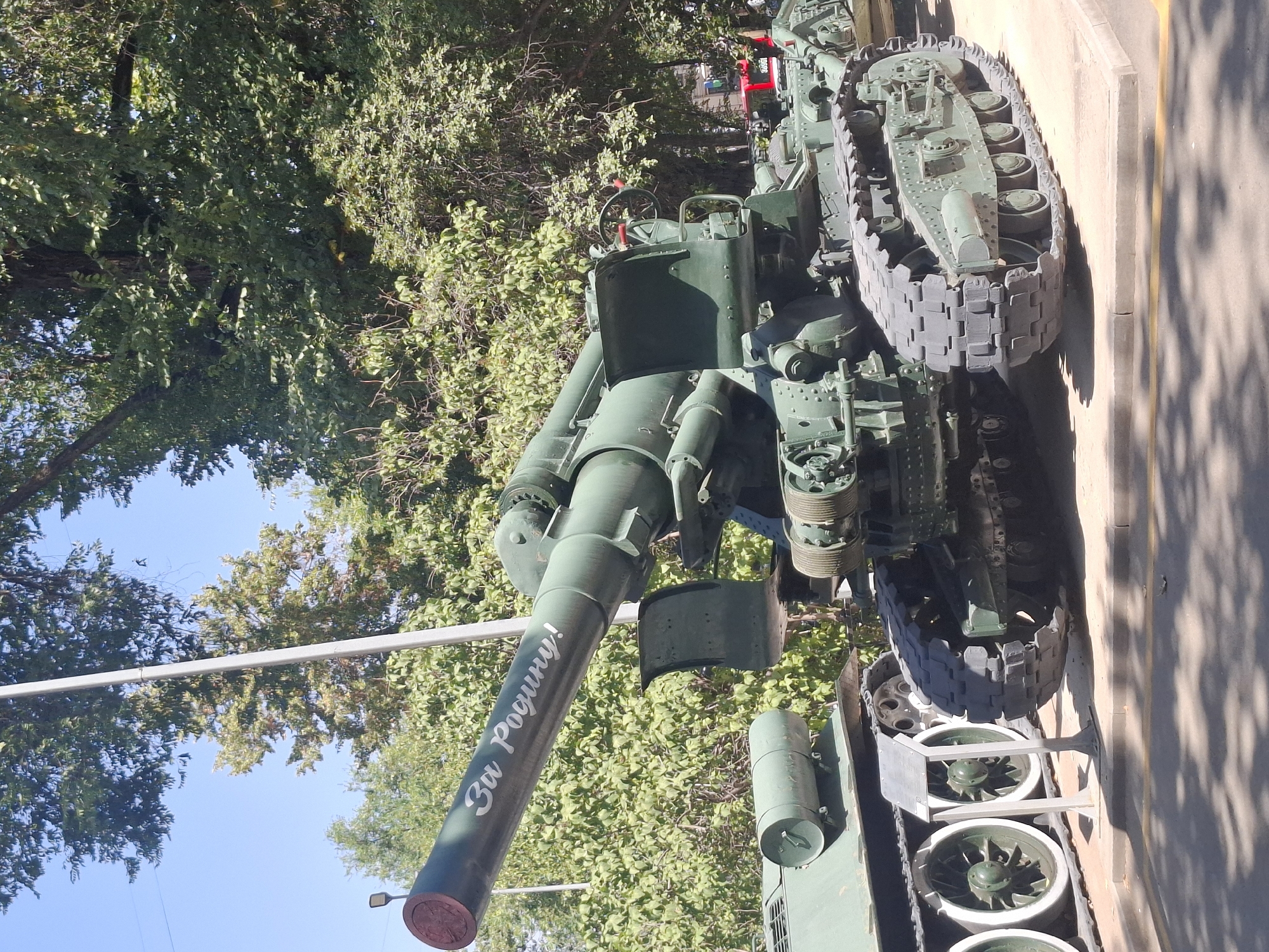 The image shows a military exhibit outdoors on a sunny day, likely in Almaty, Kazakhstan. The primary subject is a large, green, tracked artillery piece with a long barrel. The barrel has silver lettering that reads "За Родину!" (For the Motherland!). To the left of the artillery piece, the tracks and wheels of another green military vehicle are visible. The background consists of lush green trees and a clear blue sky. The artillery piece is positioned on a paved surface with a curb, and shadows from the trees are cast on the ground.