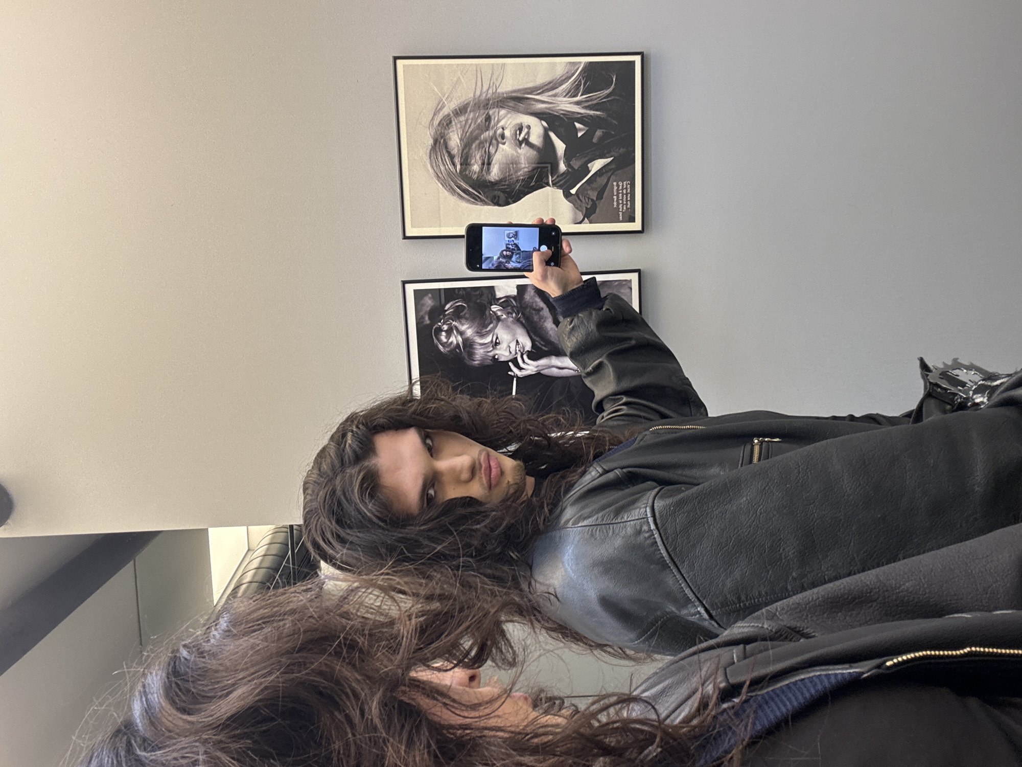 An indoor scene in Sofia, Bulgaria, features a male individual and a partial reflection on the left, likely of the same person. The male, positioned centrally, has long, dark, curly hair and wears a black leather jacket over a dark undershirt. He directly faces the viewer with a neutral, slightly pursed expression. His right hand holds a black smartphone in portrait orientation, pointing it towards a light grey wall. The phone's screen displays a distorted, internal reflection of the immediate surroundings, including portions of the wall art.
Mounted on the grey wall are two framed black and white photographic portraits of Brigitte Bardot. The left portrait depicts a smiling Bardot with a cigarette and a fur-like collar. The right portrait shows Bardot with windblown hair and a cigarette in her mouth, with French text visible below the image. The reflection on the far left shows the back and side profile of a head with long, dark, curly hair and the shoulder of a black jacket. In the upper left background, part of an industrial-style ventilation duct or pipe structure is visible near the ceiling. The overall environment is brightly lit and appears minimalist.