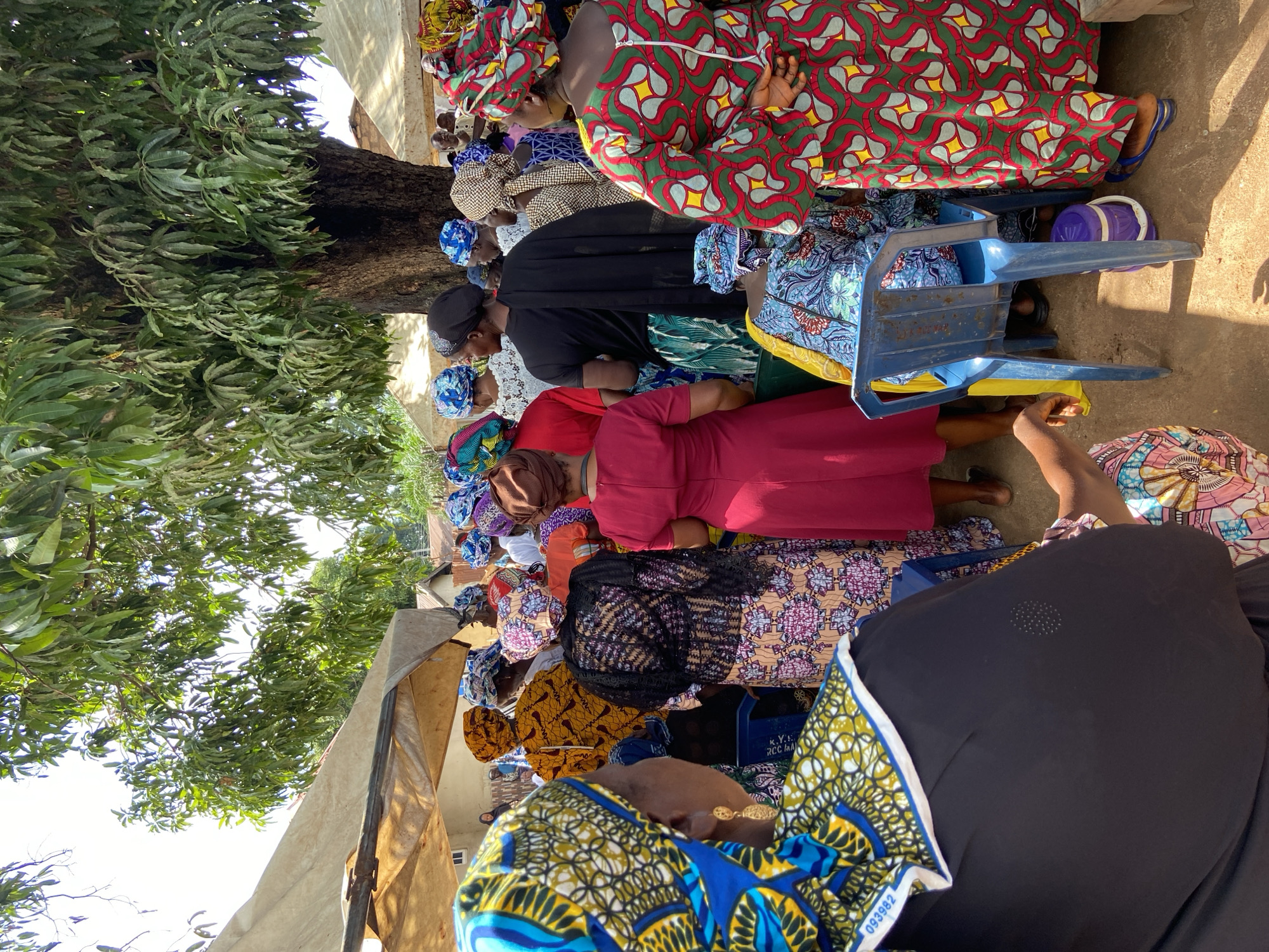 The image depicts a daytime outdoor gathering of approximately two dozen individuals, predominantly women, in Mararaba, Nigeria. Bright sunlight illuminates the scene, casting distinct shadows on the bare ground. The attendees are primarily women, many dressed in vibrant traditional African print fabrics, including patterned head wraps and full-length dresses or skirts of various designs.
In the immediate foreground, the back of a woman wearing a blue and yellow patterned head wrap, which features a white border and a visible fabric tag displaying "093982," is visible. She wears a black garment. Towards the center of the frame, a woman in a solid red dress and a brown head wrap stands near another woman wearing a red and green patterned dress with a prominent white vertical zipper down the back; her hands are clasped behind her. Other women are visible in various patterned head wraps and garments, standing in close proximity, suggesting a communal event or meeting.
The environment combines natural shade from a large, leafy tree, likely a mango tree, with artificial shade provided by light-colored tarpaulin or fabric shelters erected on frames. A blue plastic chair is present in the mid-ground, and a small purple and white insulated container rests on the ground nearby. Simple residential structures with brown roofs are visible in the distant background.