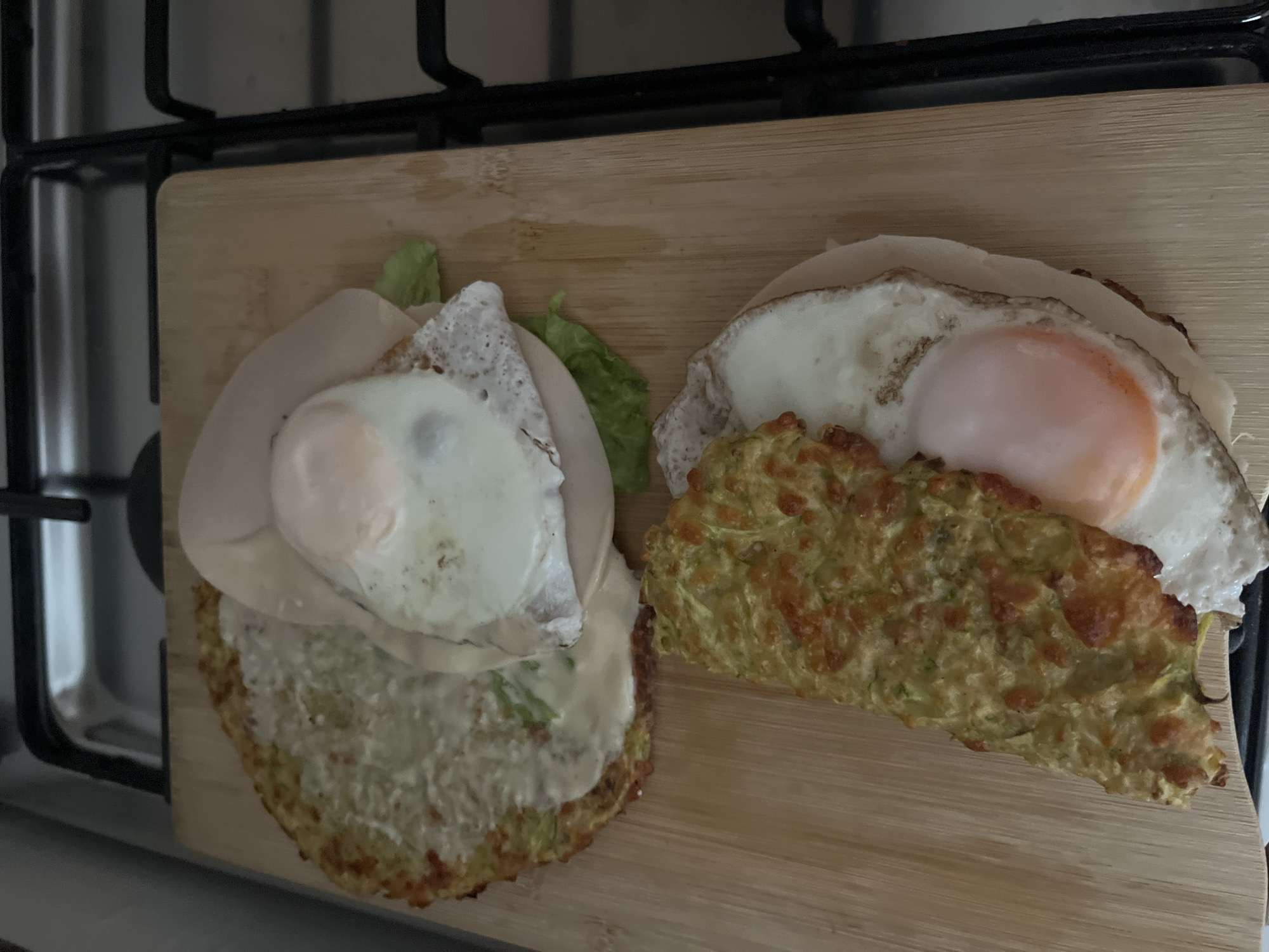 The image shows two open-faced sandwiches on a wooden cutting board, placed on a kitchen stovetop. The sandwiches appear to be made with a zucchini-based base, topped with cheese, lettuce, ham, and a fried egg. The stovetop is black with chrome accents and appears to be gas-powered. The background is blurry and out of focus, but suggests an indoor kitchen environment. There is no indication of the location being Dubai, United Arab Emirates.