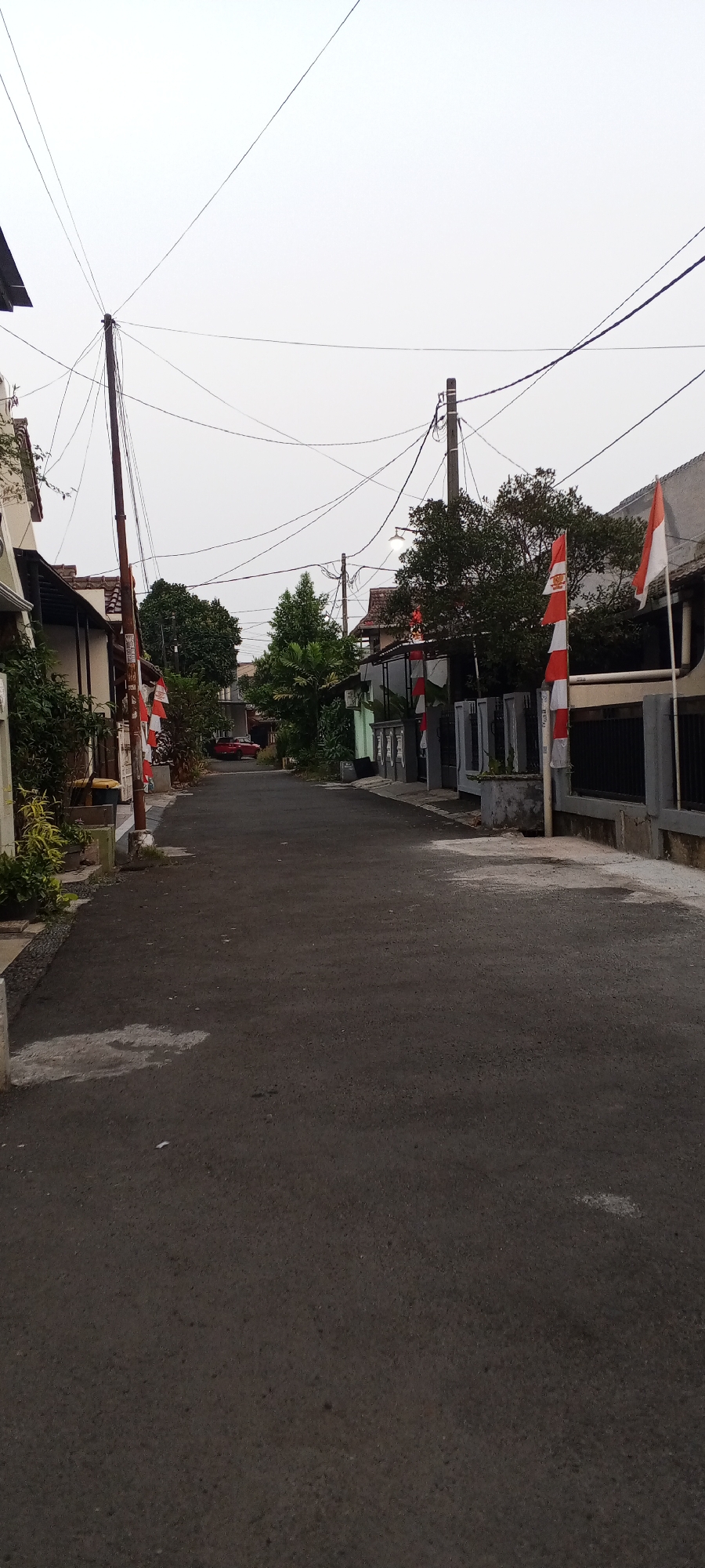 The image depicts a street in a residential area of Depok, Indonesia. The street is paved and appears to be a narrow road lined with houses and fences. Several Indonesian flags, red and white, are prominently displayed on poles along the street, suggesting a festive or patriotic occasion. Overhead, a complex network of power lines crisscrosses the sky, supported by wooden and concrete poles. Trees with dense foliage are visible in the background, some in front of houses and others forming a small cluster. A red car is parked further down the street. The sky is overcast, giving the scene a muted light. The overall impression is that of a quiet, everyday street scene in a suburban Indonesian neighborhood.