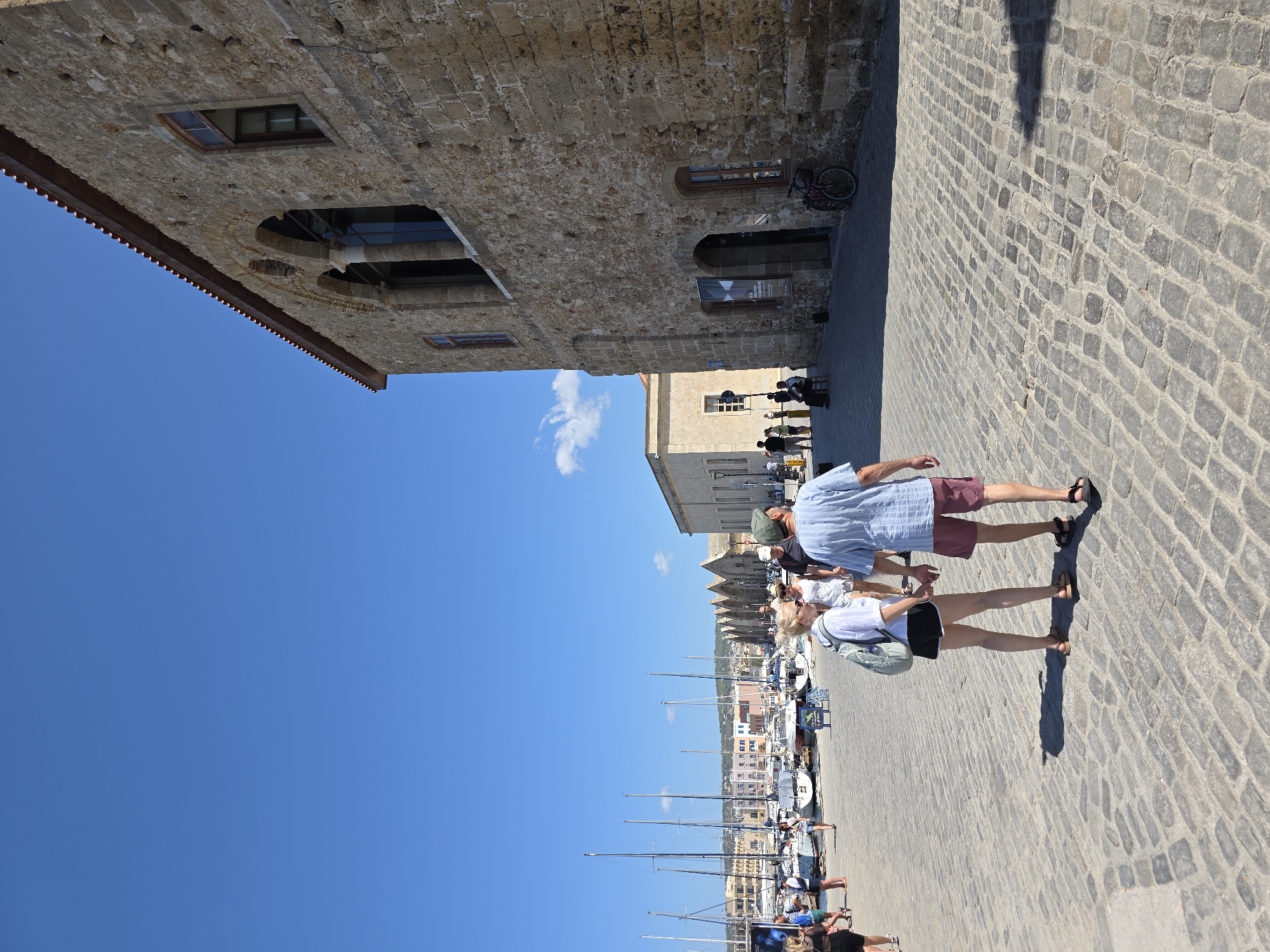 A couple, a man wearing a light blue striped short-sleeved shirt and red shorts, and a woman wearing a white short-sleeved top and black shorts, walk hand-in-hand across a cobblestone plaza. The man has a green baseball cap on. The woman has a light blue backpack. They are walking away from the camera, towards a harbor with sailboats visible in the background. To the right of the couple is a large, old stone building with arched windows. The building appears to be historical, possibly a part of Chania's Venetian harbor. The sky is clear and blue with a few wispy clouds. The scene is set in Chania, Greece, specifically in the vicinity of the Venetian Harbor, characterized by its stone architecture, cobblestone streets, and waterfront activity.