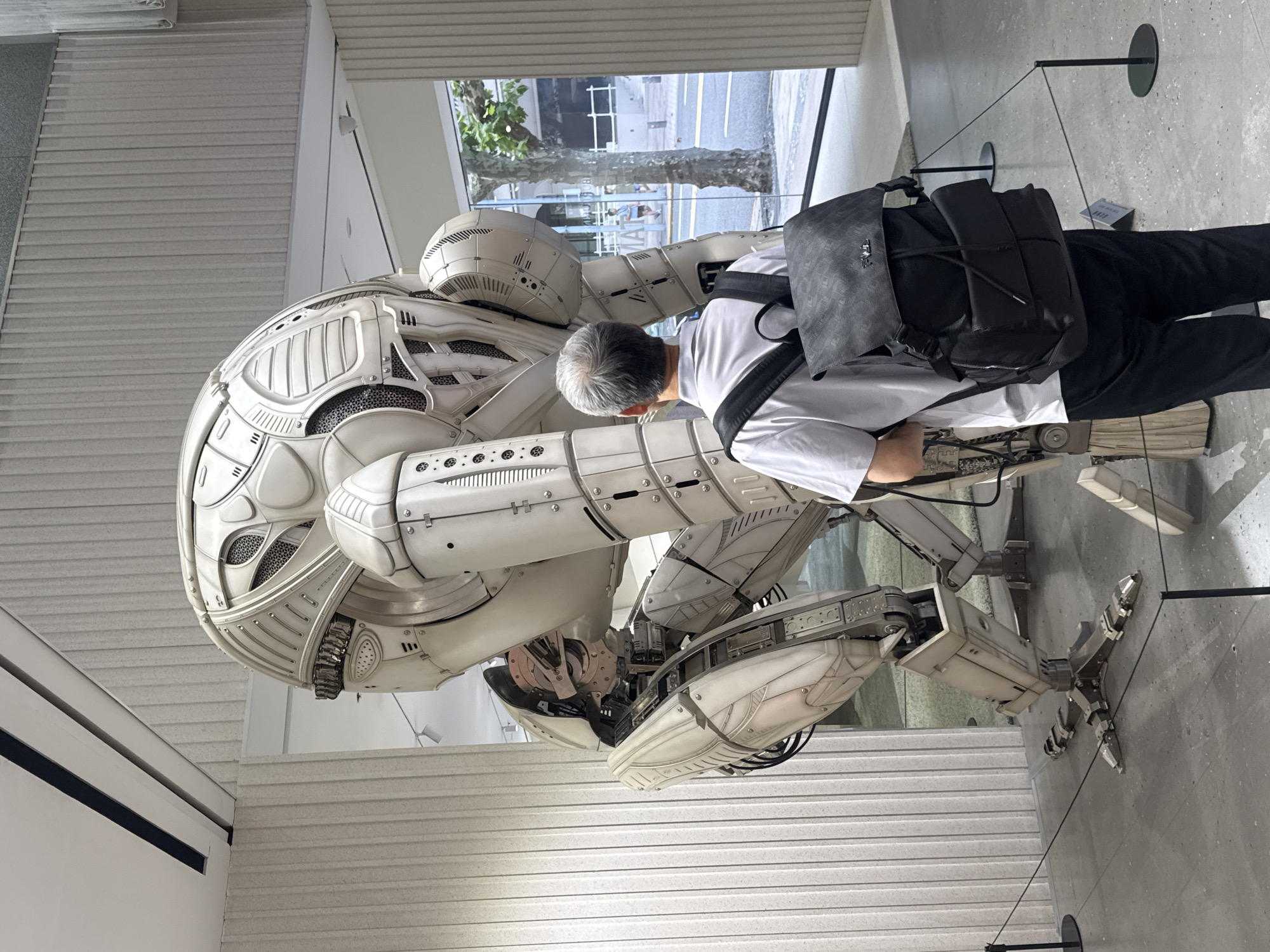A man with graying hair, wearing a white polo shirt and black pants, stands with his back to the camera, facing a large, intricate white robot sculpture. The robot has a spherical head with visible mechanical components, a segmented torso, and articulated limbs. The sculpture is positioned in a well-lit indoor space with a textured wall and a polished concrete floor. A black backpack with a textured pattern is worn by the man. Behind the sculpture, a large window overlooks a street scene with trees and buildings, indicating an urban environment. The context suggests this is a museum or gallery exhibition. The presence of a street scene visible through the window, combined with the modern architecture of the interior, points to a location in Shanghai, China, a city known for its contemporary art scene and urban development.