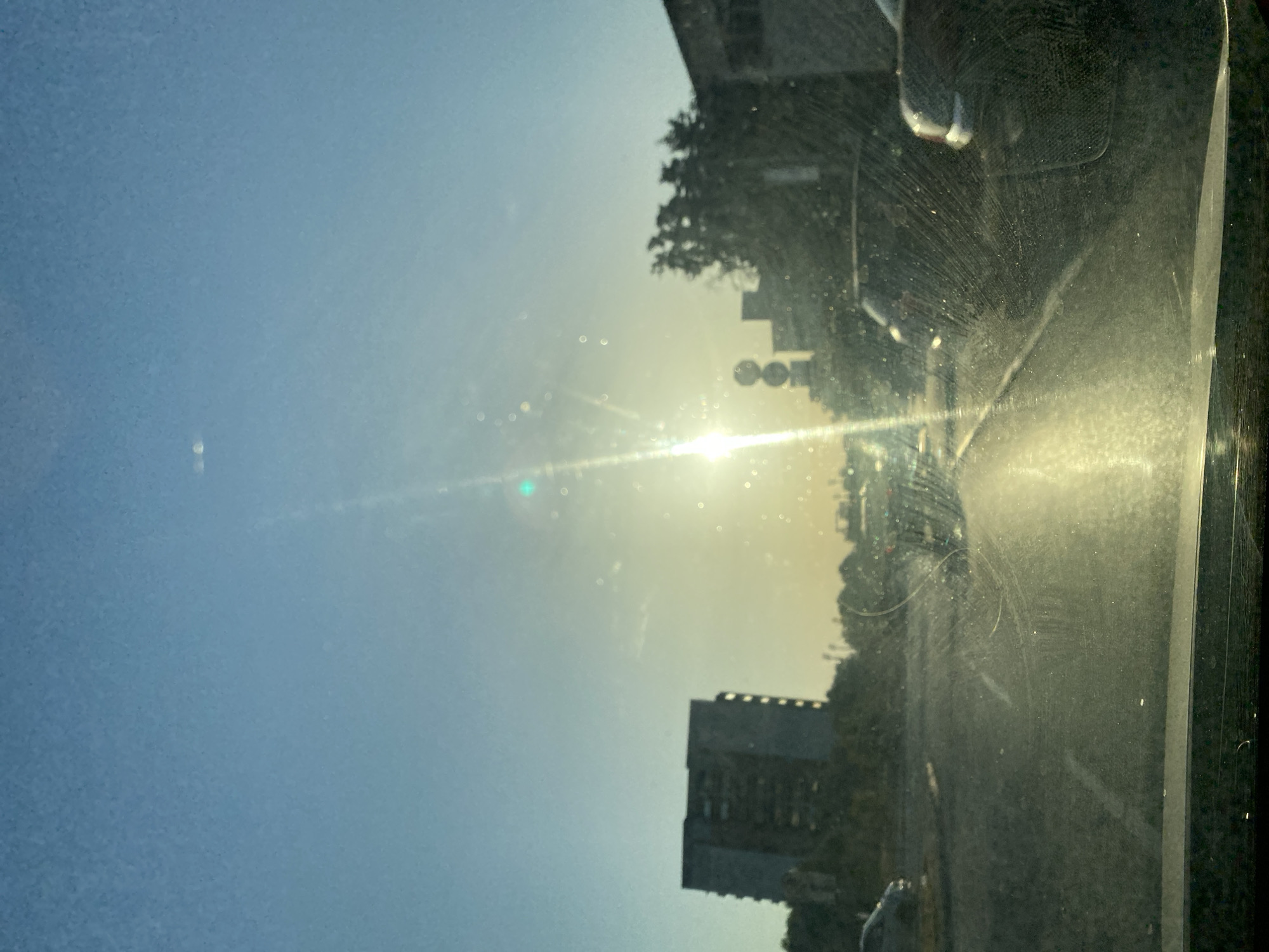 The image captures a sun-drenched road scene, likely from within a vehicle, looking out through a windshield. The sun is low in the sky, creating a bright glare and lens flares across the view. In the background, apartment buildings and trees are visible, suggesting an urban or suburban setting. The road appears to be a multi-lane street with cars driving in the same direction. The windshield itself has visible smudges and streaks, which are exacerbated by the bright sunlight. The sky is a clear blue, with the sun creating a bright, diffused light. The overall impression is one of a typical drive in a city, with the natural elements of sunlight and weather contributing to the visual effect. The location is the city of Bar, Montenegro.