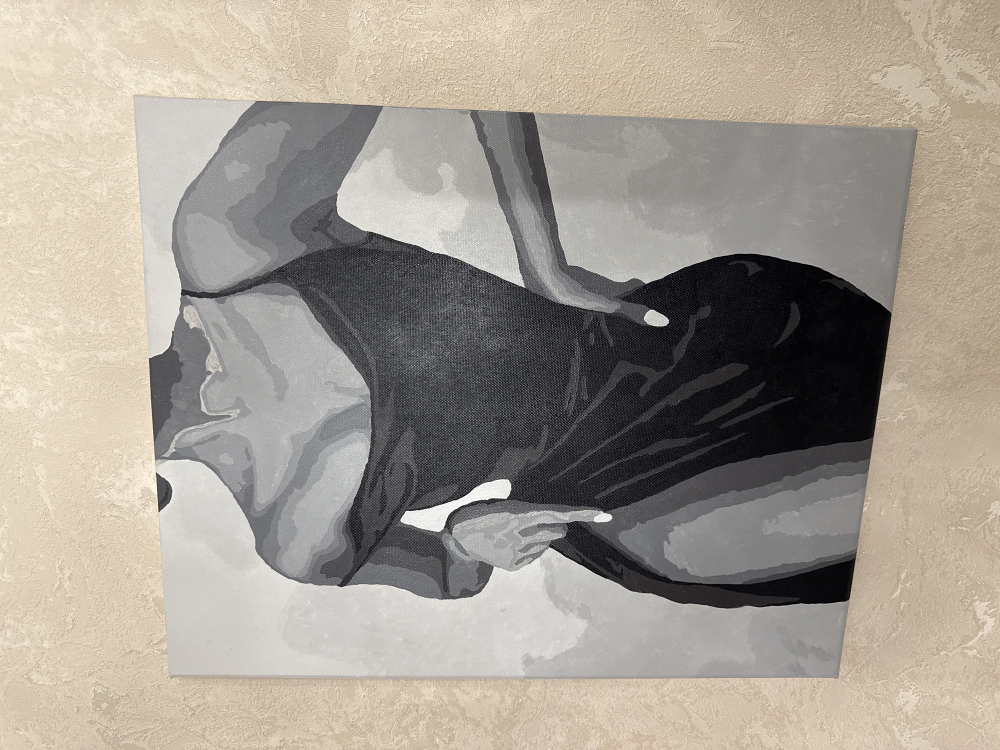 The image displays a monochromatic painting of a woman in a black dress, rendered in a graphic, blocky style. The painting is hung on a textured, cream-colored wall. The woman is depicted from the chest down, with her left hand resting on her hip and her right hand pointing downwards. The dress is strappy and appears to be made of a flowing material. The lighting in the image is soft and diffused, casting no harsh shadows. The painting is centered in the frame, and the wall texture occupies the background and the left side of the image. There is no indication of the city of Saratov, Russia, within the visual information provided by the image.
