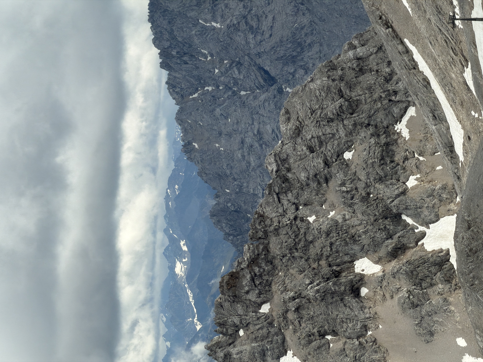 This is a landscape photograph of rugged, rocky mountains under a cloudy sky. The foreground features a steep, rocky slope with patches of snow. In the mid-ground, more jagged mountain peaks rise, also with scattered snow. The background shows a range of distant mountains, appearing bluer and softer due to atmospheric perspective, with some snow-capped peaks visible. The sky is filled with heavy, gray clouds, suggesting overcast weather. There are no people or man-made structures immediately visible in the main mountain scenery, though a thin, dark pole-like structure is present in the lower right corner of the frame, potentially part of a lift system. The scene appears to be in a high-altitude alpine environment. The location is identified as Wildermieming, Austria.