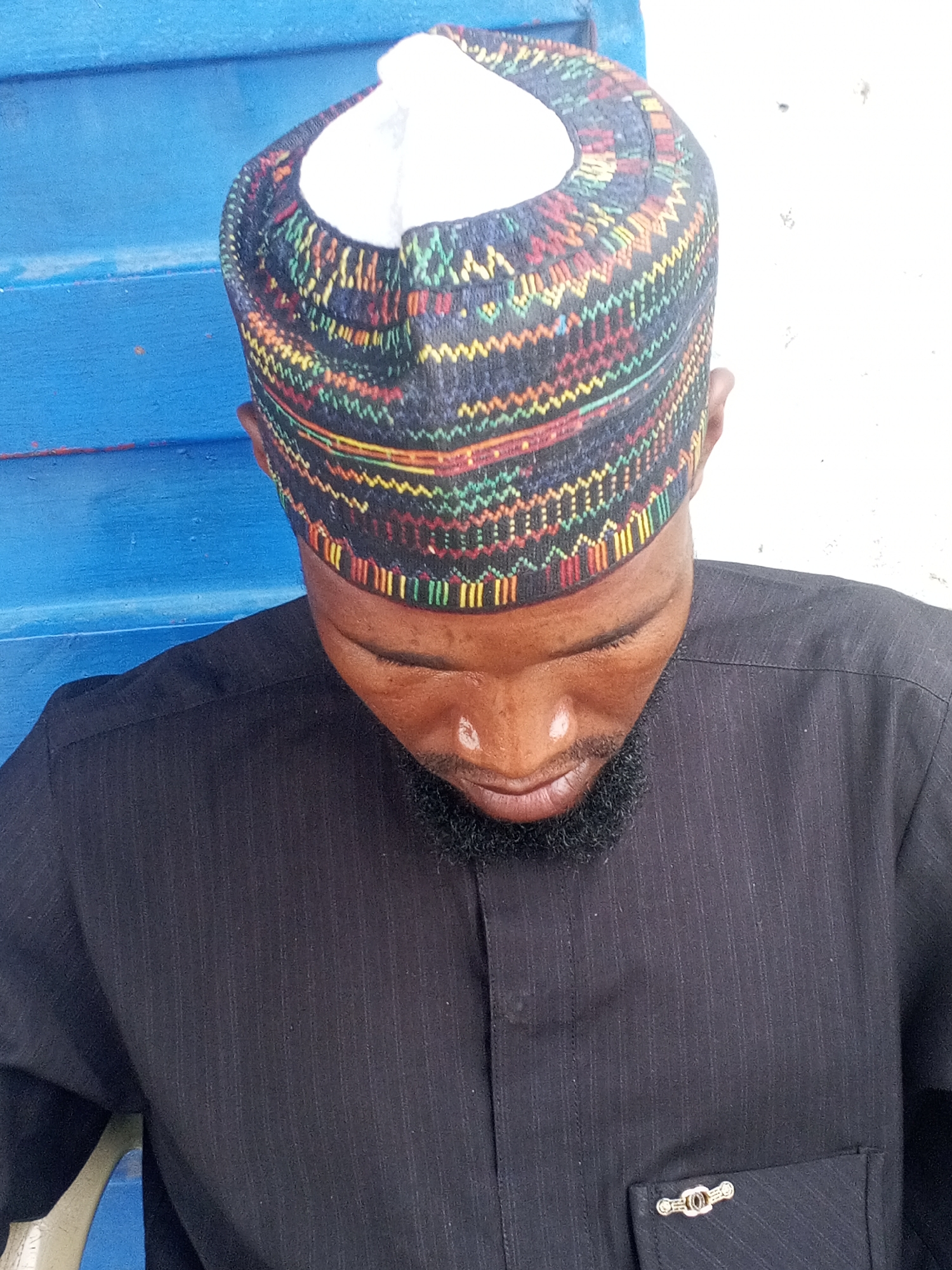 A close-up, high-angle medium shot shows a Black man wearing a black pinstripe kaftan and a colorful, patterned cap. He has a short, dark beard and appears to be looking down. The background consists of a blue wooden surface on the left and a white wall on the right. The scene is located in the city of Jos, Nigeria.