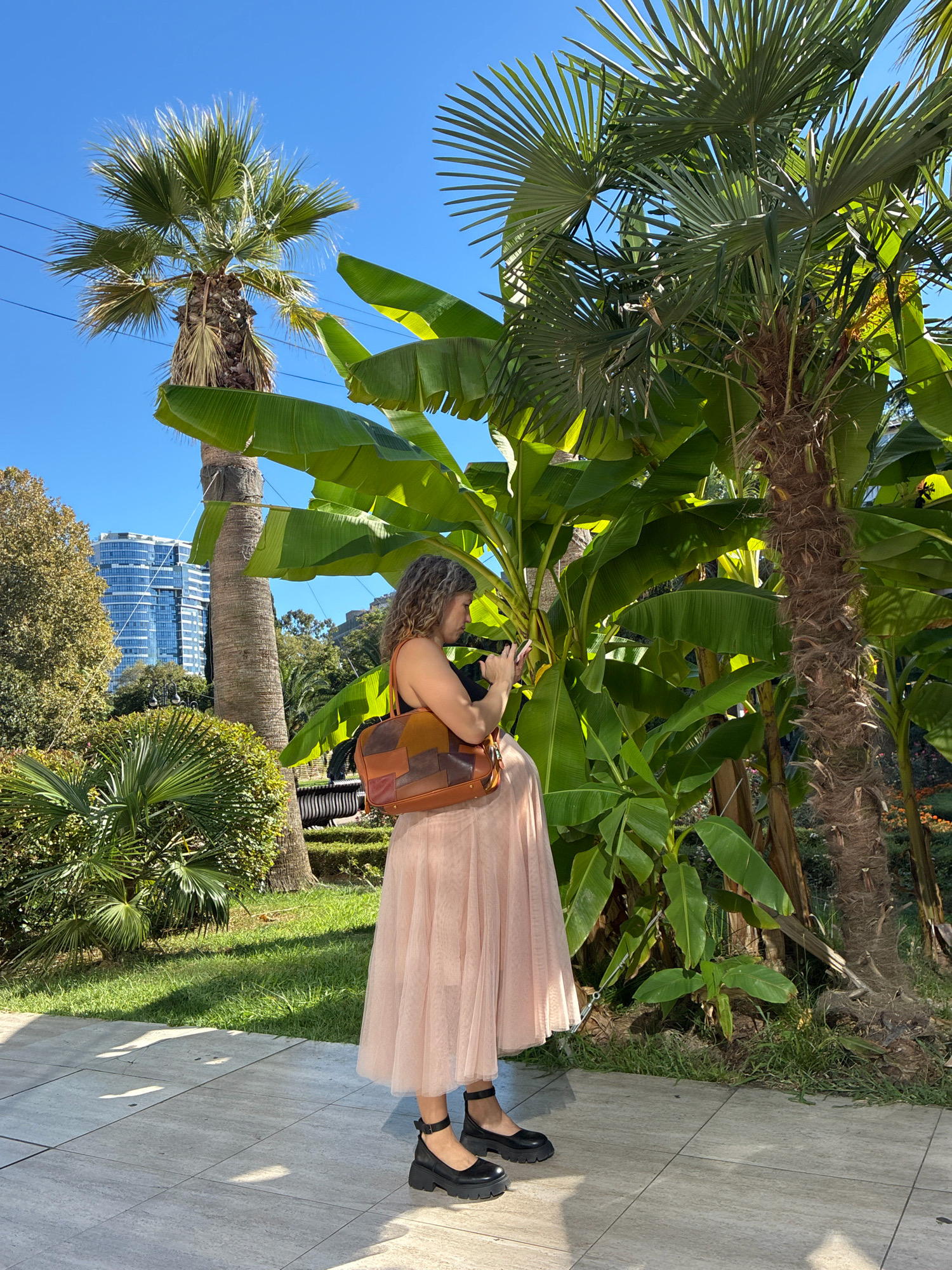 A woman stands in a park in Sochi, Russia, holding a phone and looking at it. She is wearing a pink skirt and black shoes. Behind her are palm trees and a building. The sun is shining and casting shadows on the ground.