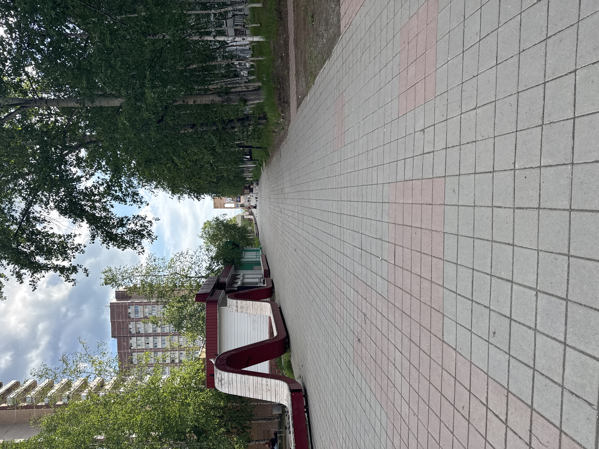 The image depicts an outdoor scene on a paved walkway in the city of Tynda, Russia. The walkway is paved with square tiles of alternating gray and pink colors. To the left of the walkway is a curved, wave-like bench structure made of wood and red metal. Behind the bench is a building with a red roof and white siding, and beyond that, a tall, multi-story apartment building with a brick facade. A line of tall trees with green foliage runs along the right side of the walkway, with birch trees prominent among them. In the distance, several people are visible walking along the path, and some pigeons are present on the walkway and the bench. The sky is cloudy. The scene suggests a public space, possibly a park or a pedestrian zone within the city.