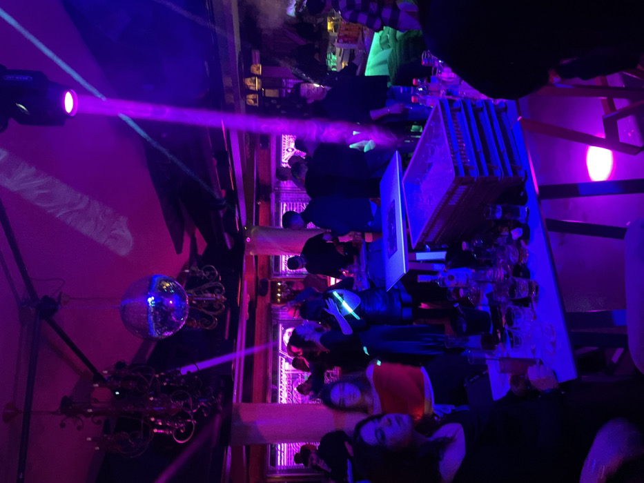 This image captures an indoor scene at what appears to be a vibrant nightclub or bar, presented with a 90-degree counter-clockwise rotation. The setting is primarily illuminated by dynamic artificial lighting, casting strong purple and blue hues across the room.
Numerous people are visible throughout the space, engaged in social interaction. Many individuals are standing, gathered around a central bar or counter area that features bottles and what look like drinking glasses. Their faces are often obscured by the dim lighting, distance, or angle, contributing to a lively, bustling atmosphere. In the foreground, closer to the viewer, a woman in a red top and another person with dark hair are discernible. One person in the mid-ground holds a bright, glowing object, possibly a glow stick.
On the left side of the scene, a prominent lighting fixture emits a powerful purple laser beam, cutting across the room. Below this, a classic disco ball hangs, reflecting ambient light. Ornate, dark-colored decorative elements, possibly chandeliers or wall sconces, are also visible near the disco ball. Some areas show a slight haze, suggesting smoke effects common in such venues.
The overall activity points to a social gathering or party during the nighttime. The venue's characteristics — special lighting, disco ball, bar setup, and crowd — are consistent with a nightlife establishment. The provided context indicates the location is Drapetsona, Greece, though no specific visual cues within the image confirm this. No discernible text is visible in the media file.