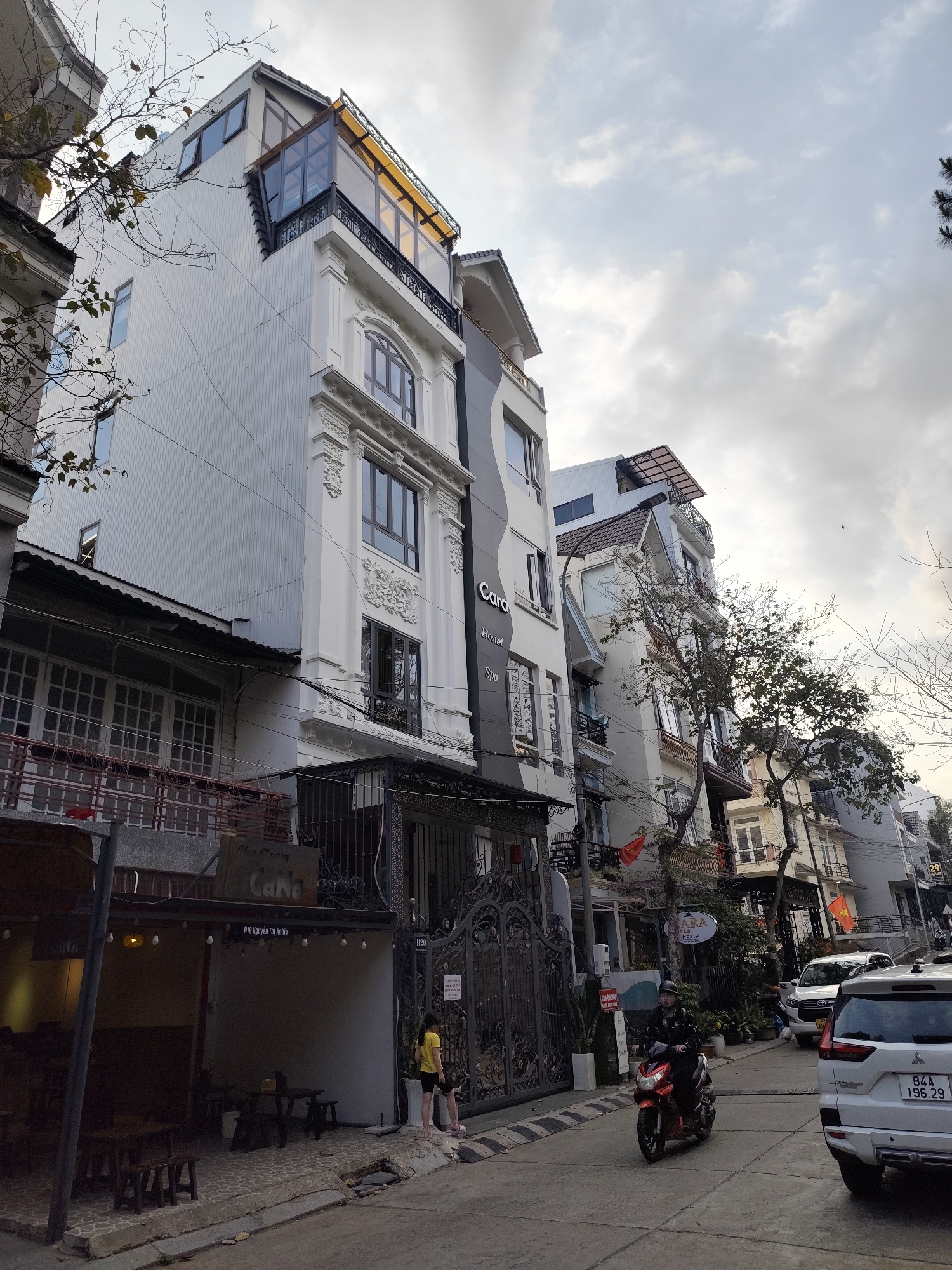 The image depicts a street scene in what appears to be a densely populated urban area of Da Lat, Vietnam. The architecture consists of a row of narrow, multi-story buildings. The building facades vary in design, some showing colonial-era decorative elements, while others appear more modern with glass-enclosed rooftop sections. Signage on several buildings indicates that they house businesses such as the "Cara Hostel Spa" and another hostel "ARA." A coffee shop called "Cha Crom CaNa" is also present.
Two people are visible; one is a female standing in the street. A person is driving a motorcycle on the road. A white SUV is positioned at the edge of the road. License plate "84A 196.29."
The street is relatively narrow, and the sky overhead shows an overcast condition. Utility wires are strung between the buildings. Bare trees and branches extend into the scene from the sides, suggesting a cooler time of year.