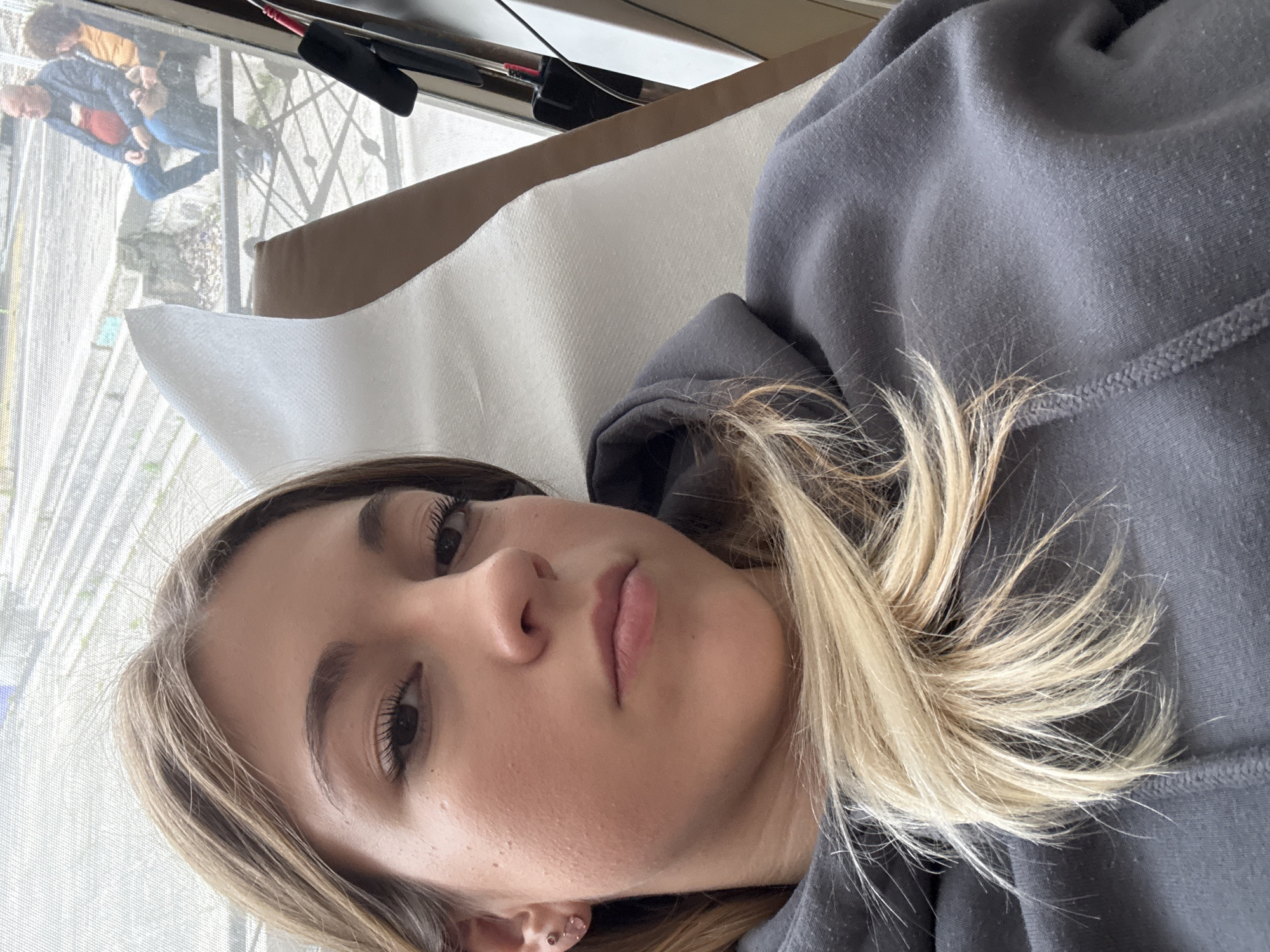 The image features a young woman captured in a close-up, seemingly reclining and looking directly at the camera with a neutral expression. She has blonde and light brown hair and is wearing a dark grey hooded sweatshirt.
Behind her, through a window covered with a fine mesh screen, an outdoor scene and other individuals are visible. Two people are identifiable: one in a yellow top and dark trousers, and another in a blue top and jeans holding a reddish object. They appear to be in a garden or courtyard with some greenery. The upper part of the window frame shows reflections or objects like a black handle with a red tip, possibly a cleaning tool or part of a window fixture. The woman is supported by a light-colored, textured pillow and a brown headrest. The lighting suggests it is daytime. The location is stated to be Sofia, Bulgaria, though no specific visual cues confirm this within the image. No text is visible.