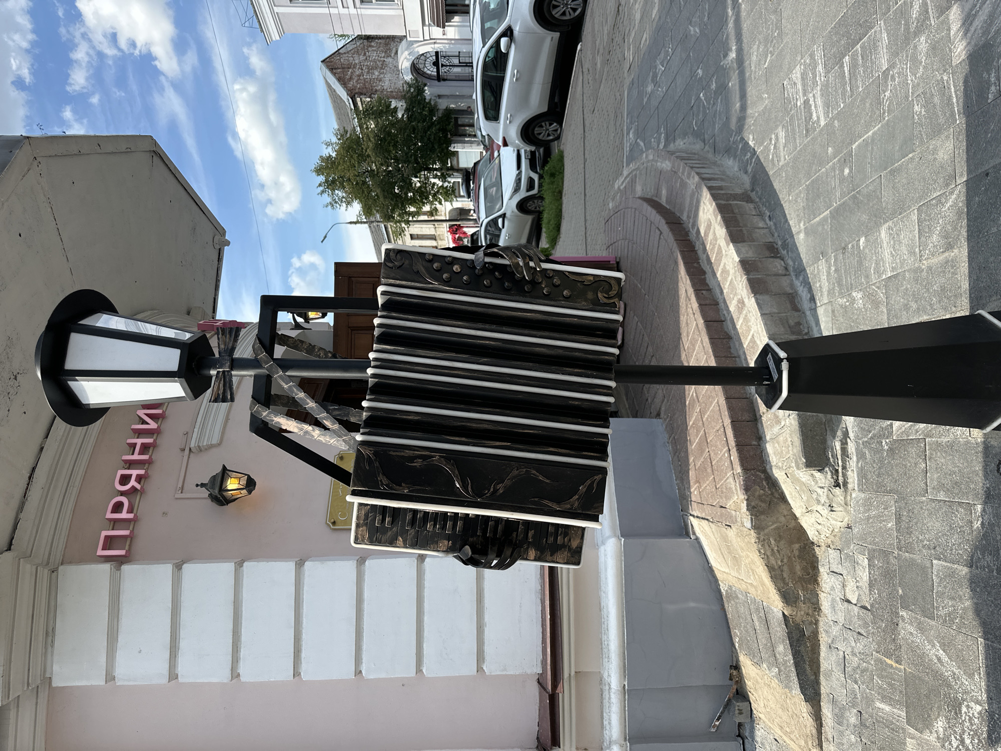 An outdoor scene in the city of Putyatino (Semivragovskaya S/A), Russia features a street with buildings and parked cars. In the foreground, a large, decorative accordion sculpture is mounted on a lamppost. The accordion is black with white keys and gold decorative elements. Behind the sculpture, a building with pink siding and white columns is visible, with a sign that reads "ПРЯНИ" in pink lettering. A vintage-style lantern hangs from the building. The street is paved with stone tiles, and several white cars are parked on the right side of the image. Trees and other buildings are visible in the background under a partly cloudy sky.