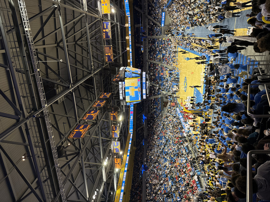 An aerial-perspective image depicts the interior of a packed sports arena, Edwin W. Pauley Pavilion, situated in Los Angeles, California. The central hardwood court, light brown with blue boundary lines, features a single visible basketball hoop. Above the court, a large, multi-sided video scoreboard displays "UCLA 11" and "N 0" as the score, with a game clock reading 11:28. The arena's ceiling structure, a complex network of metal trusses, supports numerous blue and yellow championship banners for UCLA basketball and men's volleyball, commemorating years such as 1964, 1970, 1975, 1995, and 2024. The tiered seating is largely occupied by a crowd of spectators, many wearing blue and yellow attire. In the lower seating section to the right, a university band, including a bass drum labeled 'UCLA', is visible. Individuals identified as athletes, coaches, and event staff are positioned on and around the perimeter of the court.