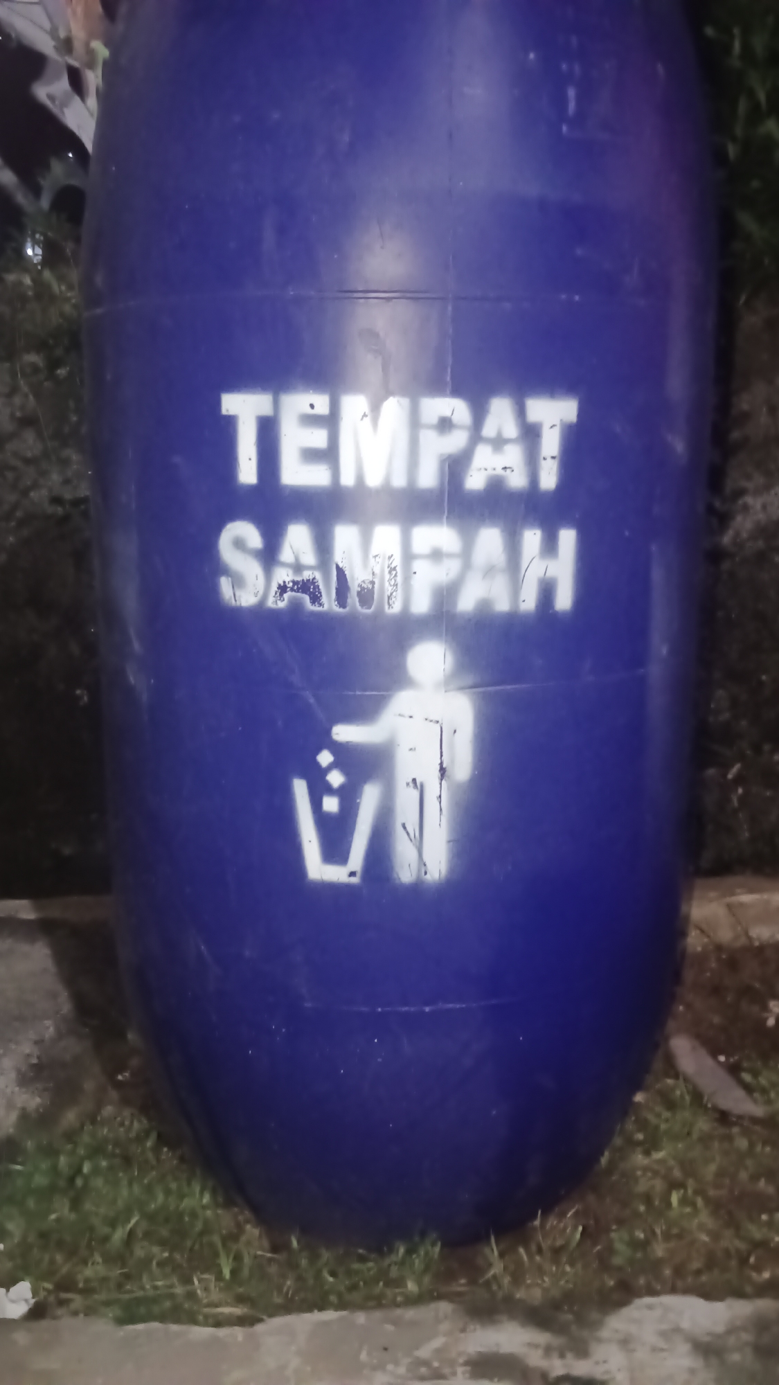 A large, blue, cylindrical trash receptacle with stenciled white text and graphics is centered in the frame. The text, in all capital letters, reads "TEMPAT SAMPAH," which translates to "Trash Can" in English. Below the text, a stencil depicts a stick figure with its arm raised, dropping items into a V-shaped receptacle, symbolizing the act of discarding waste. The receptacle appears to be made of plastic and is situated outdoors, with a grassy area and some concrete visible in the foreground. The background is dark and out of focus, suggesting the image was taken at night or in low light conditions. The overall condition of the receptacle suggests it has been used and weathered. The location is identified as Kemang, Indonesia.
