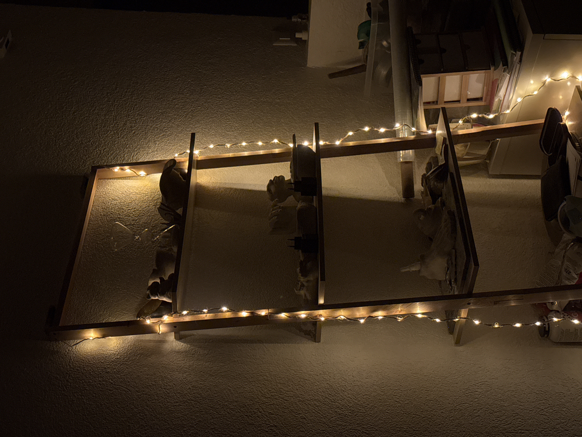 The image shows a wooden ladder shelf unit adorned with fairy lights, illuminated by a warm glow. The shelves are sparsely populated with various objects, including small sculptures or figurines, a bowl, and what appear to be art supplies or tools. The background is a textured wall, likely an interior wall of a room. The lighting suggests it is nighttime or a dimly lit interior space. No people are visible in the image. The setting is an indoor, domestic environment, possibly a studio or living space. The image does not contain any information that would allow for identification of its location within the city of Neunkirch, Switzerland.