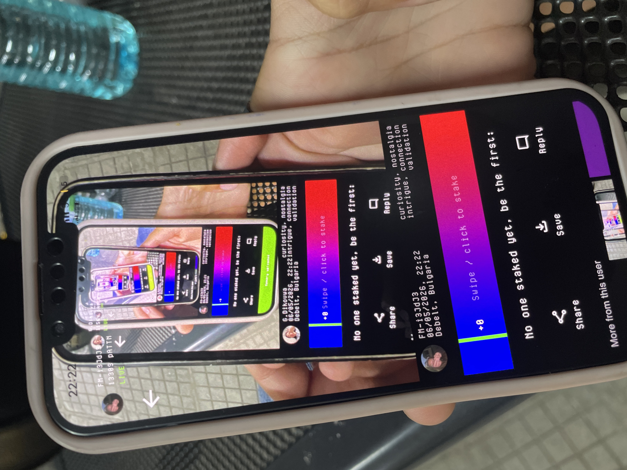 A right hand holds a white-cased smartphone vertically, displaying an application interface. The smartphone screen shows a recursively embedded image of a hand holding a smartphone, creating a Droste effect.
On the visible smartphone screen:
* The top left corner indicates the time "22:22".
* User information "G.Dikouva" and "FM-i3JdJ3" are displayed.
* A date and time "06/05/2026, 22:22" are visible, along with the location "Debelt, Bulgaria".
* Keywords such as "curiosity, nostalgia intrigue, connection validation" are also present.
* A horizontal bar featuring a blue section, a green vertical line, and a purple-to-red gradient section reads "+0 Swipe / click to stake".
* Text below this bar states, "No one staked yet, be the first:".
* Icons for "Share", "Save", and "Reply" are visible at the bottom of the visible interface.
* The nested image within the application duplicates this exact interface, including the staking bar and text.
The background includes a dark, ribbed surface on the right and a lighter, reflective surface on the left. A blurry, translucent blue plastic bottle containing liquid is present in the upper right background. The general environment appears to be an indoor setting.