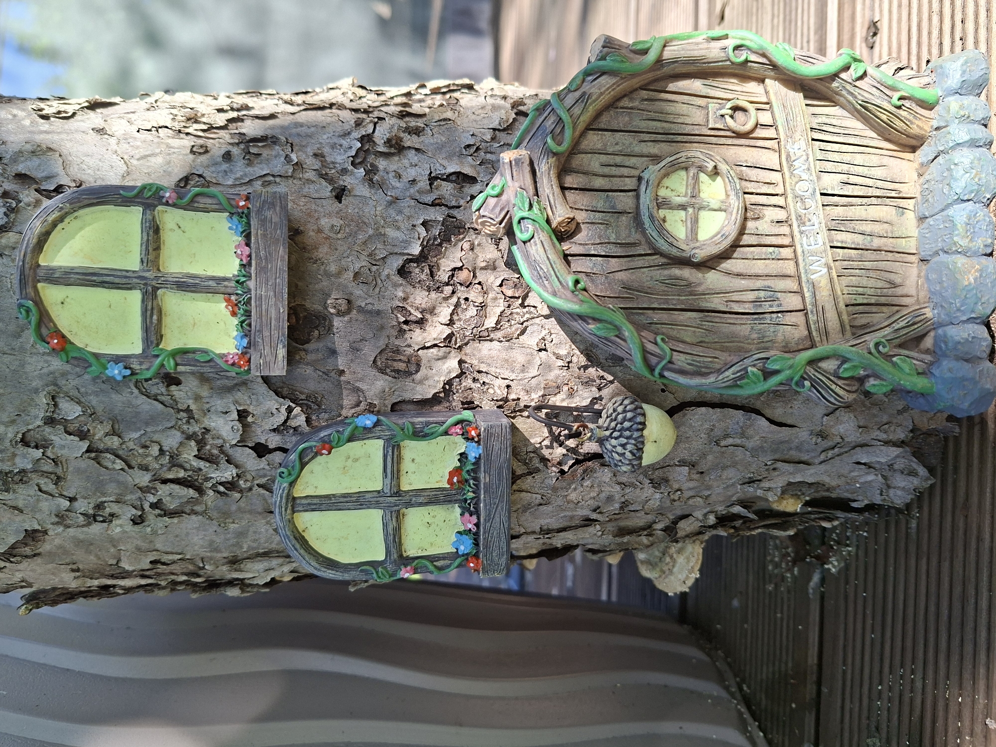 This image depicts a whimsical display of miniature fairy house elements attached to a large tree trunk, likely in a garden setting within Vansi, Estonia. The tree has rough, textured bark. Mounted on the trunk are two small, arched windows with green ivy accents and tiny colorful flowers along the sills. Below these windows, a larger, arched fairy door is prominently displayed. The door is made to resemble wood grain and features a small round window at the top, a ring-shaped handle, and the word "WELCOME" embossed across the middle. Green vines further adorn the door. Hanging from the tree trunk, near the door, is a decorative acorn. The background shows a blurred outdoor environment with wooden decking visible at the bottom of the frame.