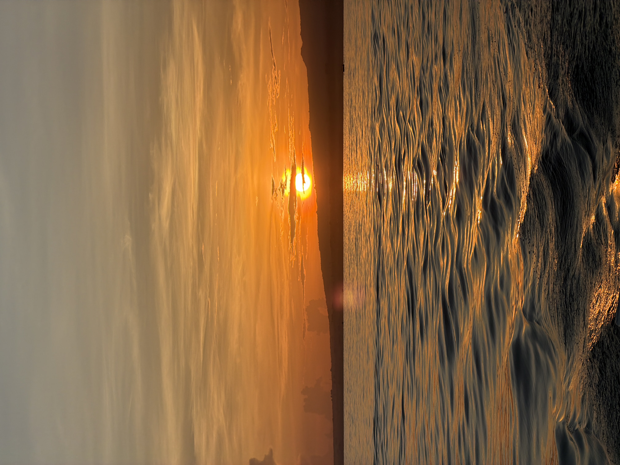 This image depicts a sunset over the ocean in Temukus, Indonesia. The sun is setting over the ocean, with its golden light reflecting on the water and the sand. The sky is a mix of orange, yellow, and grey, with clouds scattered throughout. The ocean is relatively calm, with gentle waves lapping at the shore. The beach is visible in the foreground, with the wet sand reflecting the sunlight. The image appears to be split vertically into two different sections: on the left, the sunset sky, and on the right, the ocean and beach. There are no people or other objects visible in the image.