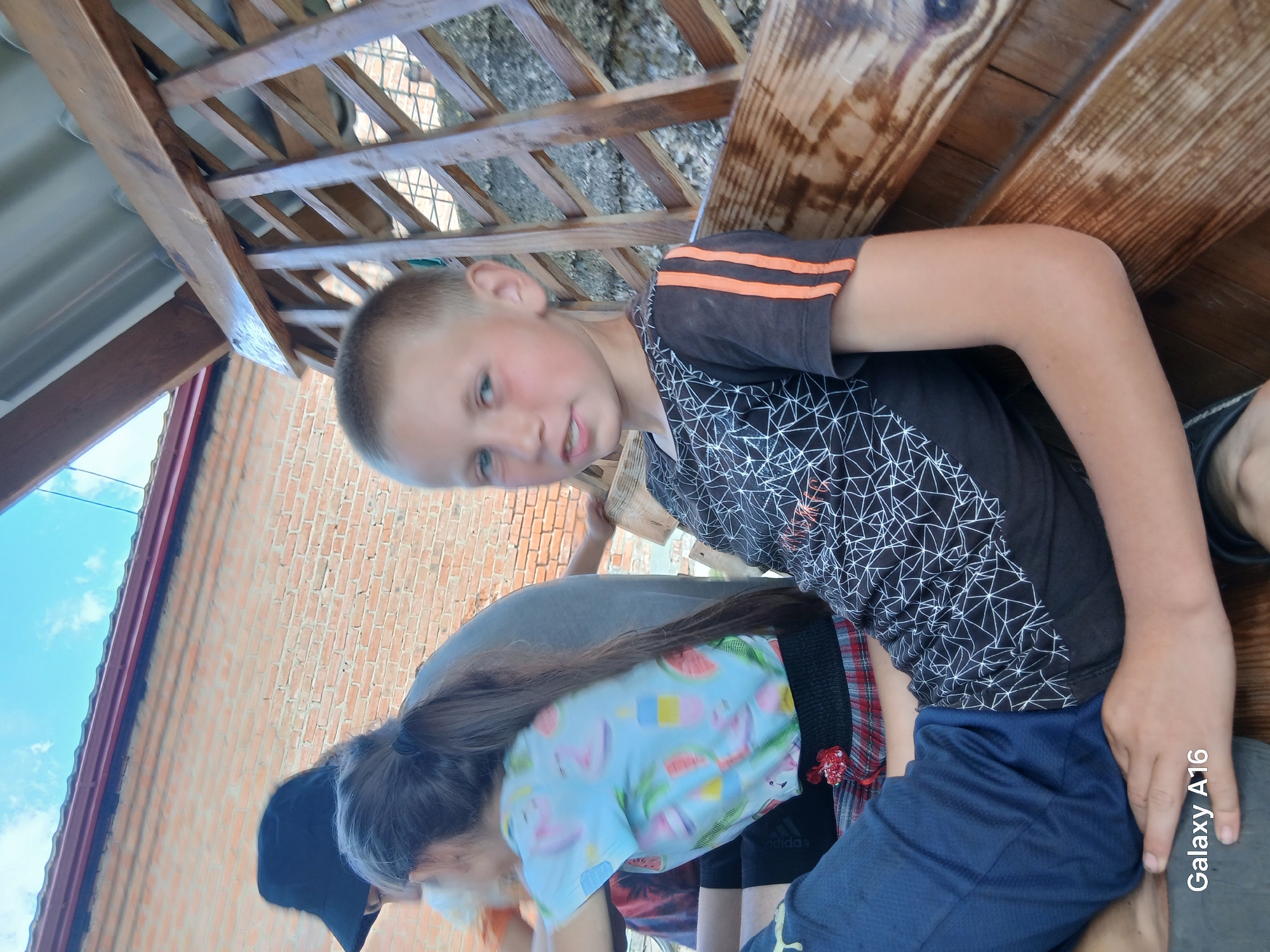 This media file shows a group of people, likely children, gathered under a wooden structure, possibly a gazebo or porch. A young boy in a black t-shirt with orange stripes on the sleeves is in the foreground, looking towards the camera with a slight smile. His skin is fair, and he has short, light-colored hair. Behind him, another person, likely a girl, is wearing a light blue t-shirt with a pattern of watermelons and popsicles. She has long, dark hair and is wearing a black hat. The background features a brick wall, suggesting a rural or suburban setting. The overall impression is one of a casual, outdoor gathering. The lighting suggests it is daytime, with the sun casting shadows and illuminating the scene. Visible text includes "Galaxy A16" on the right side of the image, indicating the device used to capture the photo. The location is Ustyvytsia, Ukraine.