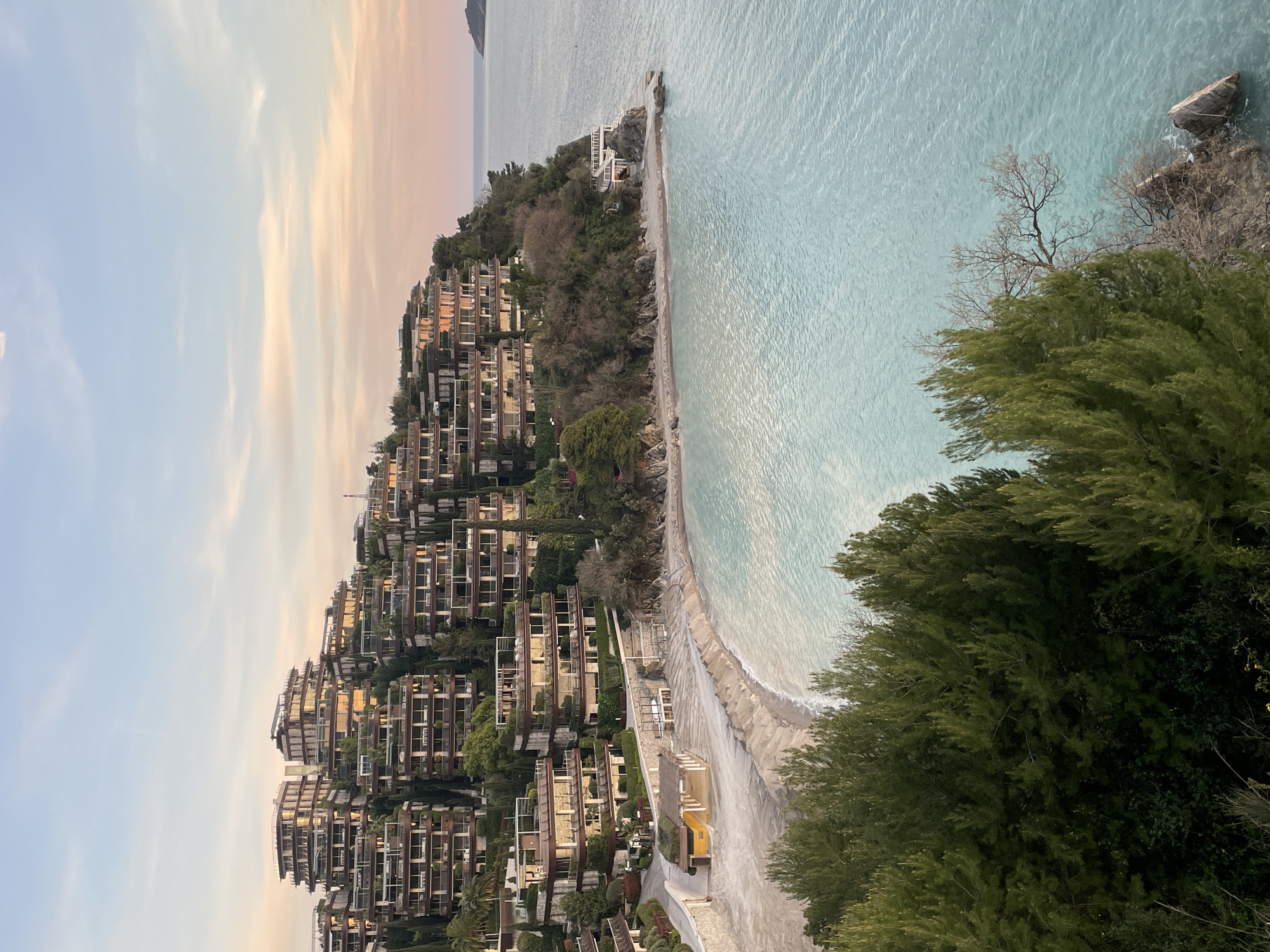 An elevated, wide-angle image depicts a coastal resort complex on the Zavala Peninsula in Budva, Montenegro, at twilight or early evening. The scene is dominated by numerous multi-story, terraced buildings constructed into a lush, green hillside on the left and center. These modern structures feature extensive glass facades and large balconies, reflecting the warm hues of the sky. Dense evergreen trees and manicured vegetation are interspersed among the buildings and cover the surrounding terrain. Below the buildings, a crescent-shaped sandy beach, characterized by light beige sand, meets a calm, clear turquoise sea. On the beach, a structured area includes a building with a dark roof, an adjacent shaded structure, and rows of stacked lounge chairs. Further right, another white, multi-level structure with a balcony is built into a rocky outcrop at the water's edge. The sky above transitions from light blue to soft pink and orange near the horizon on the right, where a small, dark island is visible in the distance. The foreground features the tops of dense evergreen trees. No human figures or discernible activity are present.