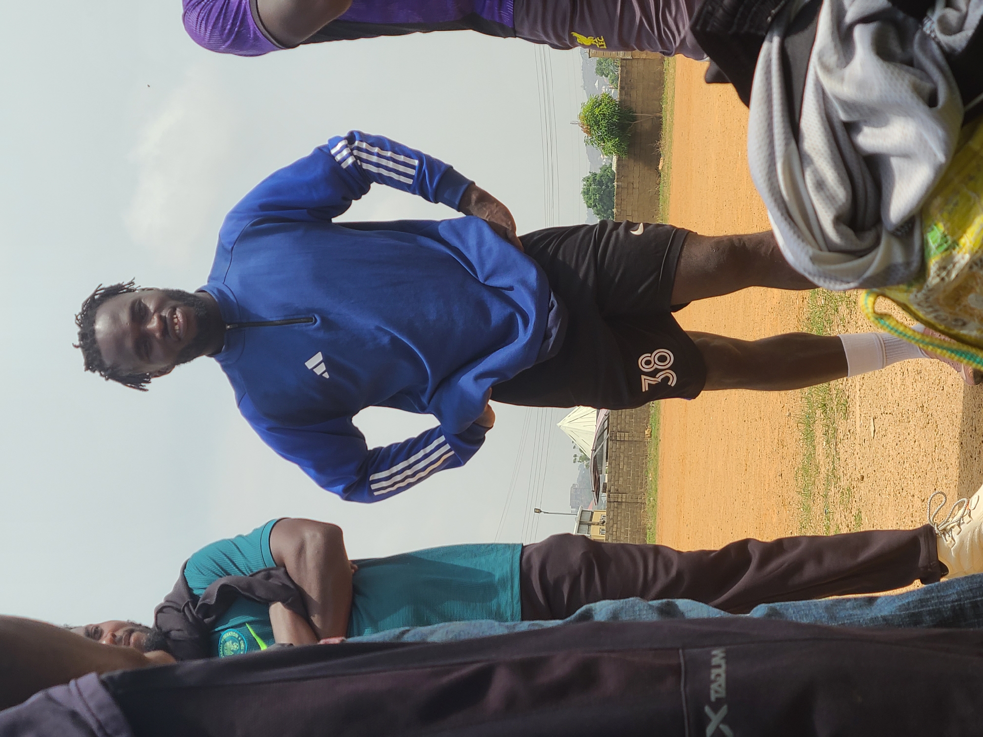 A group of people are on a dirt field. One man is centered in the frame wearing a blue Adidas sweatshirt and black Nike shorts with the number 38 printed on the thigh. Other people, wearing athletic clothing, surround him. In the background, there is a low brick wall, green vegetation, and buildings. This scene is located in Bukuru, Nigeria.