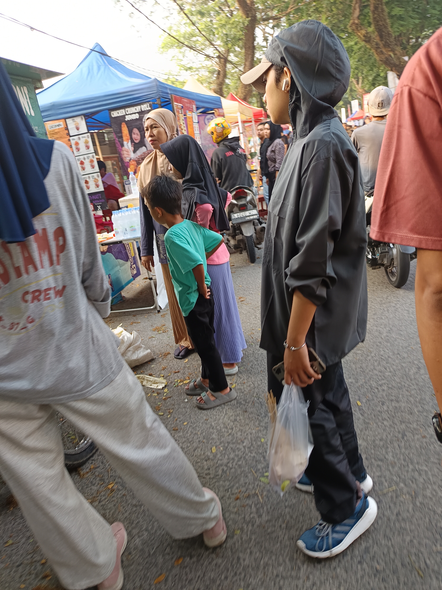 The image depicts a street scene, likely a market or a gathering, in Sodong, Indonesia. In the foreground, a person wearing a grey hooded jacket and a baseball cap walks forward, holding a plastic bag and a smartphone in their right hand. Their left arm is bent, and they are wearing a bracelet. To their left, a person in a grey long-sleeved shirt with "STAMP CREW" printed on it walks away from the camera, their legs clad in light grey sweatpants and pink sneakers. Partially visible to the left of the person in the grey hooded jacket is a stand with a blue tent. Behind the person in the grey hooded jacket, a young boy in a turquoise t-shirt and black pants stands with his hands behind his back. To the boy's left, two women are standing, one in a beige hijab and the other in a black hijab. Further back, a motorcycle with a yellow helmet on it is visible. The ground is paved and scattered with fallen leaves. In the background, there are trees and more stalls.