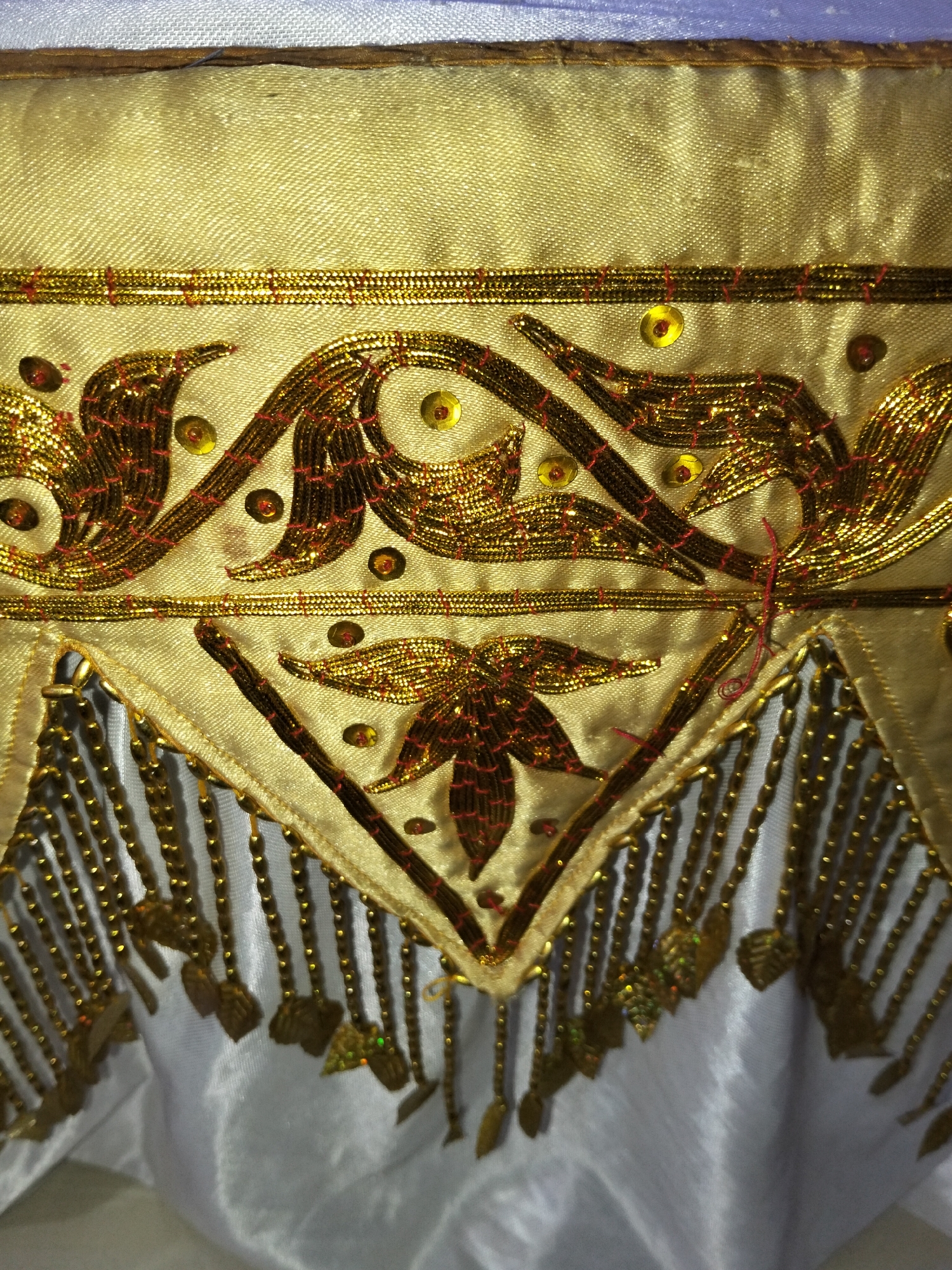 The image is a close-up of an ornate textile, likely a garment or decorative fabric, featuring intricate gold embroidery and beadwork. The primary fabric appears to be a pale gold or beige silk with a visible weave. A horizontal band of layered gold metallic trim runs across the upper portion of the image, above a decorative motif. This motif consists of swirling, curvilinear patterns embroidered in gold thread, interspersed with small, reddish-brown sequins and tiny gold sequins. Beneath this upper motif, a triangular section of the pale gold fabric is bordered by gold trim. From the lower edge of this triangle hang numerous strands of gold beads, each tipped with a small, leaf-shaped gold embellishment. The textile below this beaded fringe is a smooth, white or light gray fabric, likely satin, which drapes loosely. The lighting in the image is somewhat harsh, creating highlights and shadows that emphasize the textures and dimensionality of the embroidery. There are no people or environmental elements visible beyond the textile itself, and no specific location in the city of Cottrueng, Indonesia can be determined from the image.