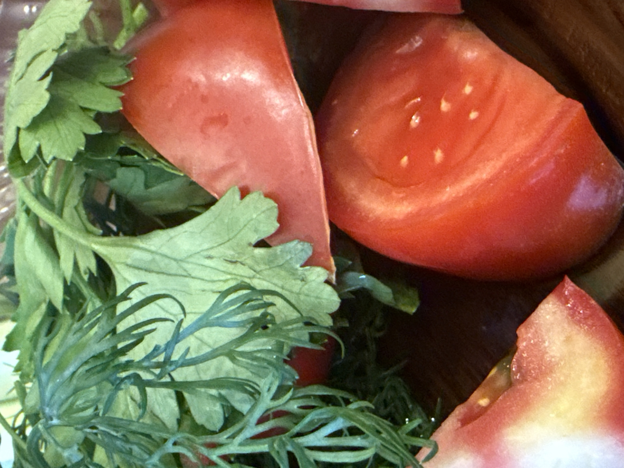 The image shows a close-up of chopped tomatoes and fresh herbs. The tomatoes are bright red and appear to be ripe. They are cut into wedges, revealing the juicy interior with visible seeds. Interspersed among the tomatoes are sprigs of green herbs, likely parsley and dill, with their finely cut leaves. The scene is tightly cropped, focusing solely on the food items, and the lighting suggests an indoor setting, possibly a kitchen or dining table. The overall impression is one of freshness and preparation for a meal, without any specific event or time of day indicated. There is no visible text in the image.