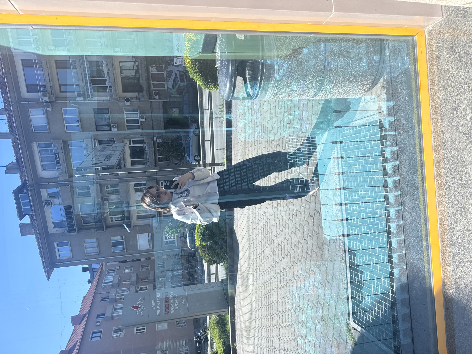 The image shows a reflection of a street scene in a window. In the reflection, a young woman with dark hair is visible, holding a smartphone and seemingly taking a selfie. The background of the reflection includes a building with multiple windows and air conditioning units, a street with a vehicle parked on it, and street-level landscaping.
The location is Sofia, Bulgaria, as indicated by the sign in the lower-left portion of the reflection, which has Bulgarian text. The sign contains a logo featuring a bird in red and white, possibly marking a business or institution. The window itself has a yellow frame and reflects a paved area with a metal grid pattern. Also visible is a dark trash can and a section of wall.