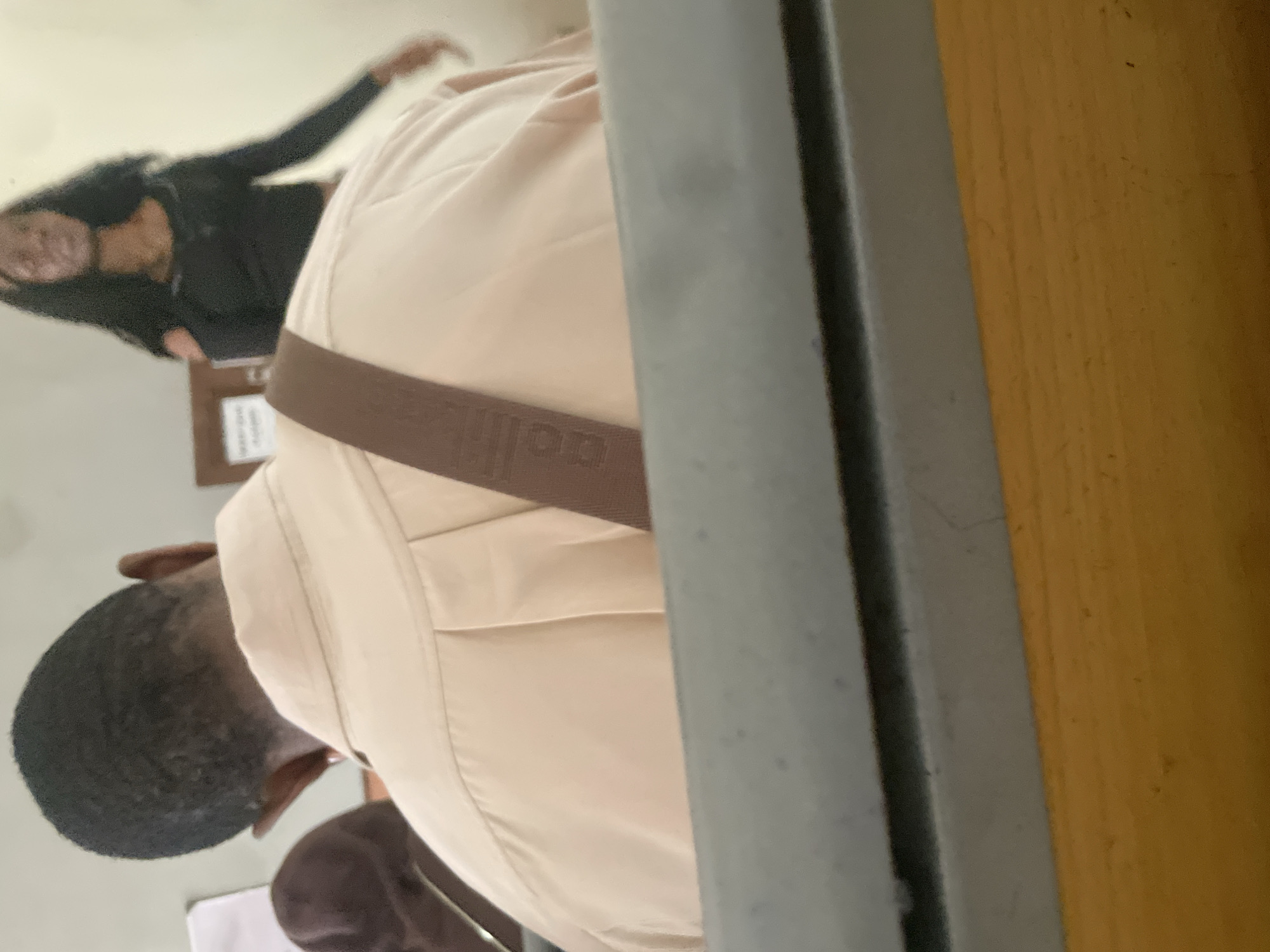 The image shows a back view of a Black person wearing a light-colored collared shirt with a brown strap across their back. The person has dark, short hair. In the background, another Black person, a woman, is visible wearing a black top and long dark hair. They appear to be in a room with a light-colored wall and some wooden furniture. The scene suggests an indoor setting, possibly a classroom or lecture hall, given the presence of desks or seating in the foreground, which are out of focus and appear to be made of wood and possibly metal. There are some papers visible on a wall or bulletin board behind the people. The overall lighting is soft. The image appears to have been taken in Ihiagwa, Nigeria.