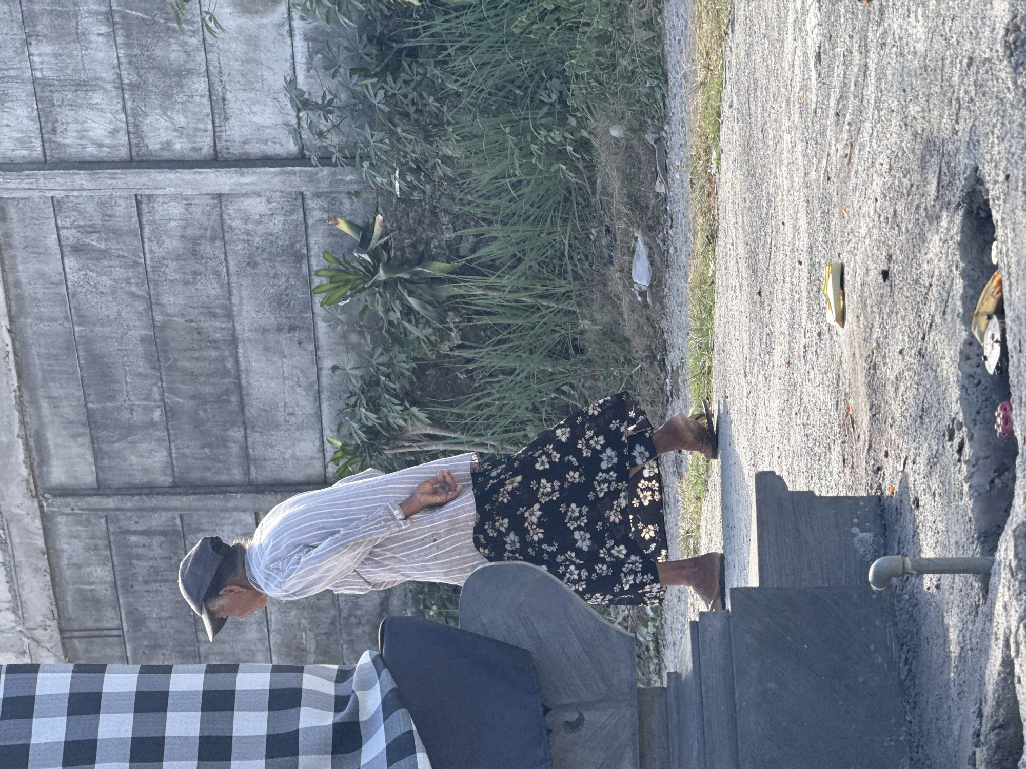In Kuta Selatan, Indonesia, an elderly woman walks past a stone structure on a sun-drenched, outdoor path. She is wearing a striped, light-colored button-up shirt and a dark, floral-patterned skirt, along with sandals. Her hands are clasped behind her back. The stone structure, to her left, is partly obscured by a large checkered cloth and features decorative carvings and a small metal pipe emerging from the ground in the foreground. The background consists of a concrete wall, a patch of dry grass, and some green foliage. Litter, including a yellow item and possibly some discarded food or offerings, is scattered on the ground and in a small opening in the path.