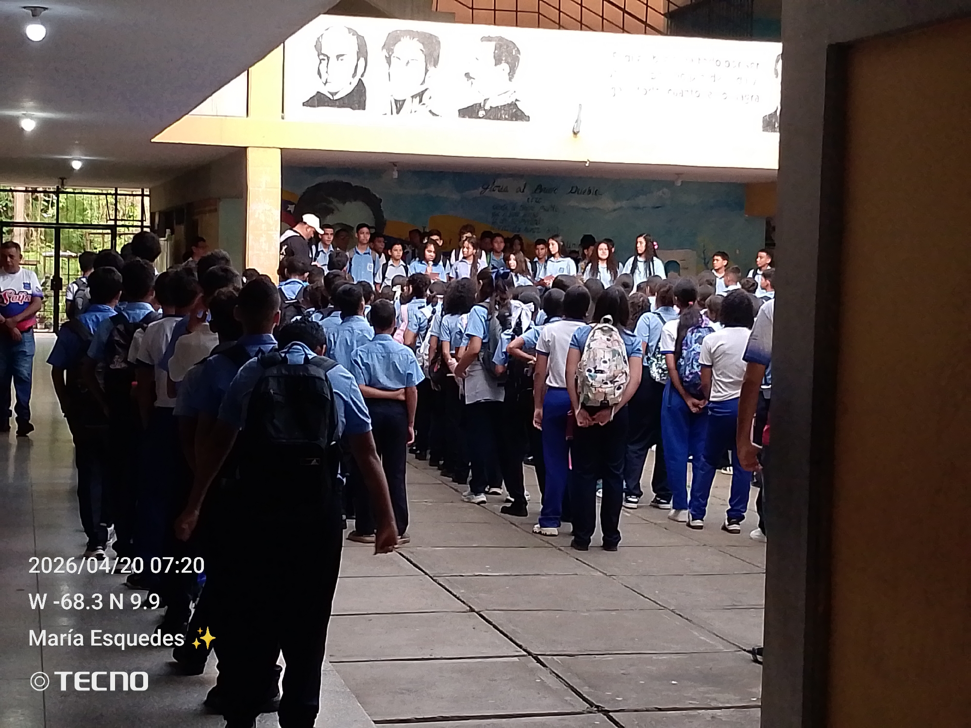 The media file displays an outdoor or semi-outdoor scene, likely within a school courtyard or covered corridor in Tinaquillo, Venezuela. A large group of approximately 40-50 individuals, primarily students, are gathered, most wearing light blue uniform shirts and dark trousers or skirts. Many students have backpacks, suggesting a school environment. They appear to be standing in organized lines or groups, facing towards an unseen point, indicative of an assembly or morning ceremony.
In the background, a building wall is adorned with a prominent mural. The upper part of the mural features three black-and-white portraits of historical figures, with the leftmost figure resembling Simón Bolívar. Below these portraits, a larger, blue-toned mural includes the phrase "Gloria al Bravo Pueblo," which is the title of Venezuela's national anthem. Other less legible text is also present on this mural. The ceiling above is simple, with exposed light fixtures. To the far left, an adult male in a white and blue patterned shirt stands observing the students.
Notable details captured in the image include a timestamp "2026/04/20 07:20," indicating an early morning shot. Geolocation coordinates "W -68.3 N 9.9" are displayed, along with the name "María Esquedes" and the camera brand "TECNO." The atmosphere appears orderly and composed, reflecting a typical school gathering.