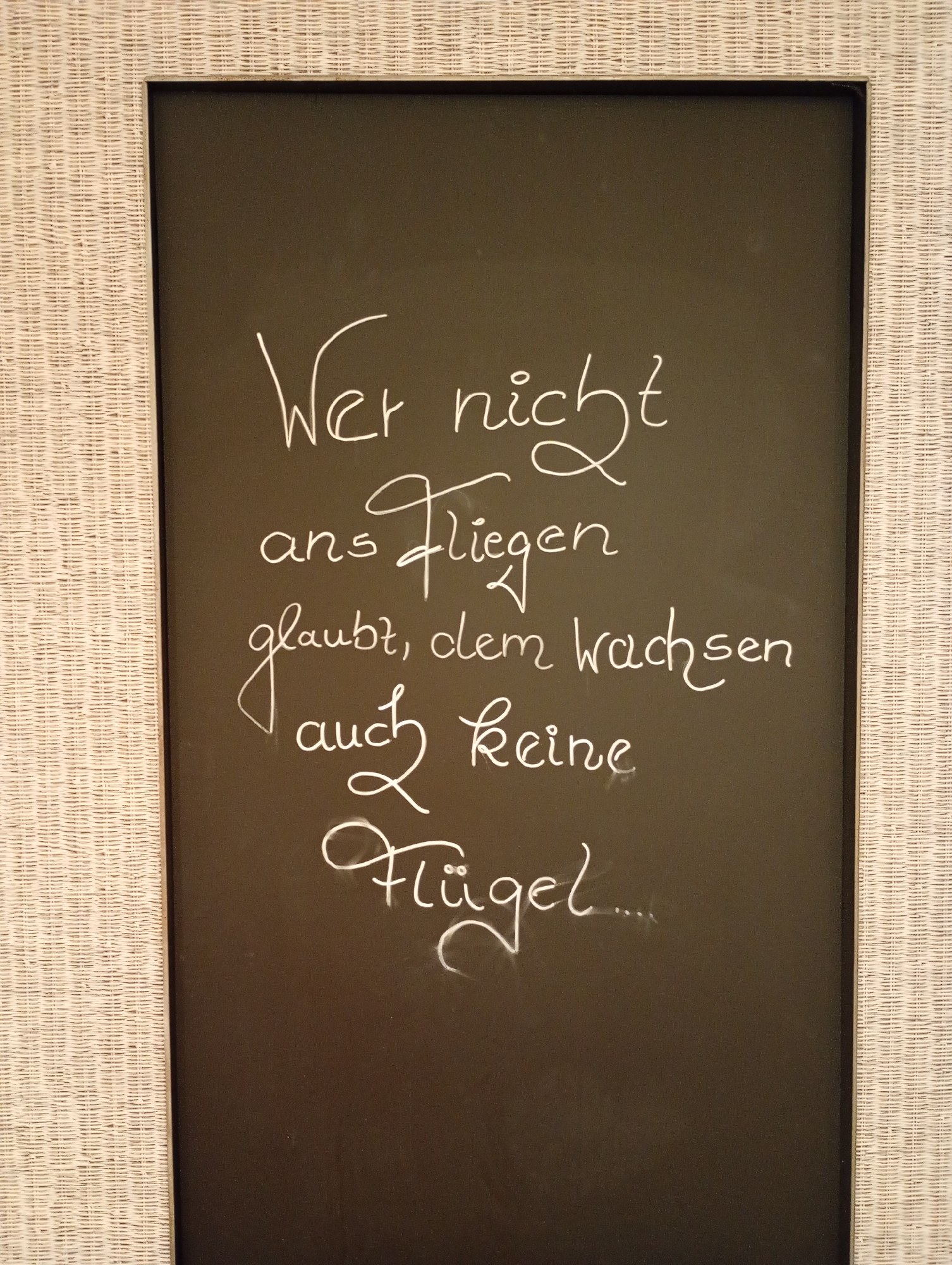 This is a photograph of a blackboard with white chalk lettering inside a woven white frame. The blackboard is dark and appears to be smooth. The lettering is handwritten in a cursive style and reads: "Wer nicht ans Fliegen glaubt, dem wachsen auch keine Flügel...". This translates from German to English as: "He who does not believe in flying, also grows no wings...". The blackboard is positioned vertically. The frame surrounds the blackboard on all four sides, with the woven texture being the most prominent feature of the frame. The background behind the blackboard is not visible. The photograph captures the blackboard at a slightly angled perspective, with the light source appearing to be from the top left, casting faint shadows. This object is likely a decorative item or a sign. Without further context, its specific location within Bad Sachsa, Germany cannot be determined.