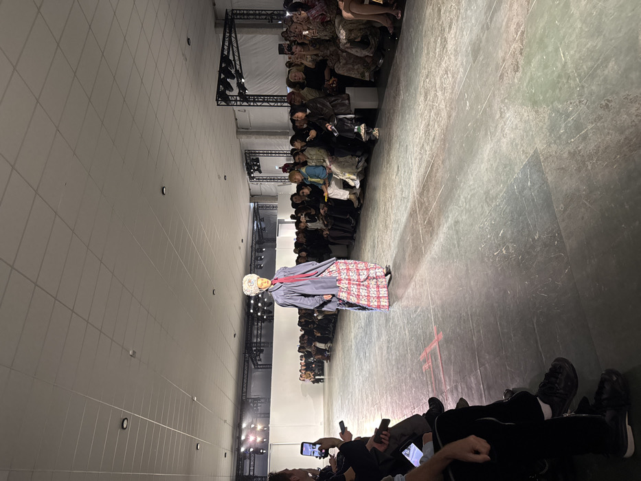 This image depicts an indoor fashion show taking place in Paris, France. A single model is centrally positioned on a grey, textured runway, walking from left to right. The model is clad in an elaborate ensemble featuring a large, ornate, predominantly white and multicolored headpiece, a loose-fitting dark blue or grey long-sleeved top with a prominent red necktie or scarf, and a long, flowing skirt or wide trousers adorned with a red and white patterned design. The model's left foot is advanced.
On both sides of the runway, an audience is seated on multiple tiers. Numerous individuals are visible, predominantly observing the model, with many holding up smartphones to record or photograph the event. The audience on the left appears closer to the camera, with a person's upper torso and arm visible in the foreground, also holding a smartphone. The general attire of the audience is varied, leaning towards dark colors, though lighter shades and patterns are also present.
The venue features a white tiled false ceiling with recessed lighting. The runway is illuminated by bright overhead spotlights, casting shadows and highlights. Behind the audience, white backdrops are visible, and further back, black truss structures with stage lighting equipment are present, indicating a professional event setup. Red tape markings in an 'X' pattern are visible on the concrete-like floor of the runway to the left of the model.