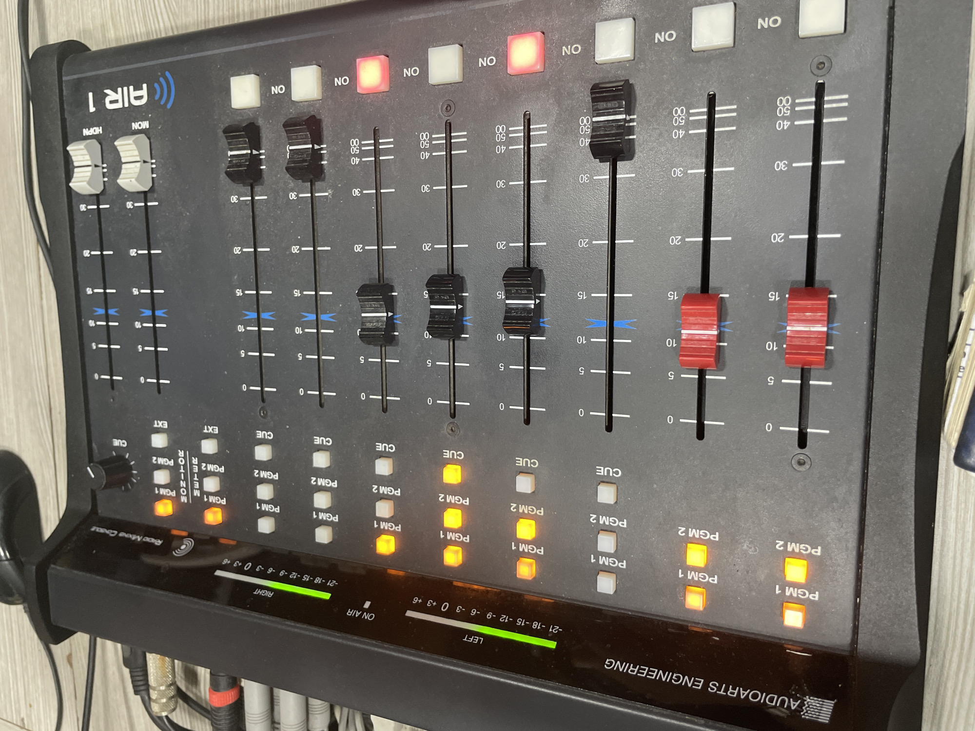The image shows an Audioarts Engineering Air 1 radio mixing console. The console has multiple channels, each with a fader, cue buttons, program buttons (PGM1 and PGM2), and channel on/off buttons. Several PGM1 and PGM2 buttons are illuminated. The monitor section is visible with a rotary knob and EXT buttons. The level meters at the bottom show a green signal. Various audio cables are connected to the back of the console. The background is a light-colored surface. There is no indication of this image being in Porlamar, Venezuela.