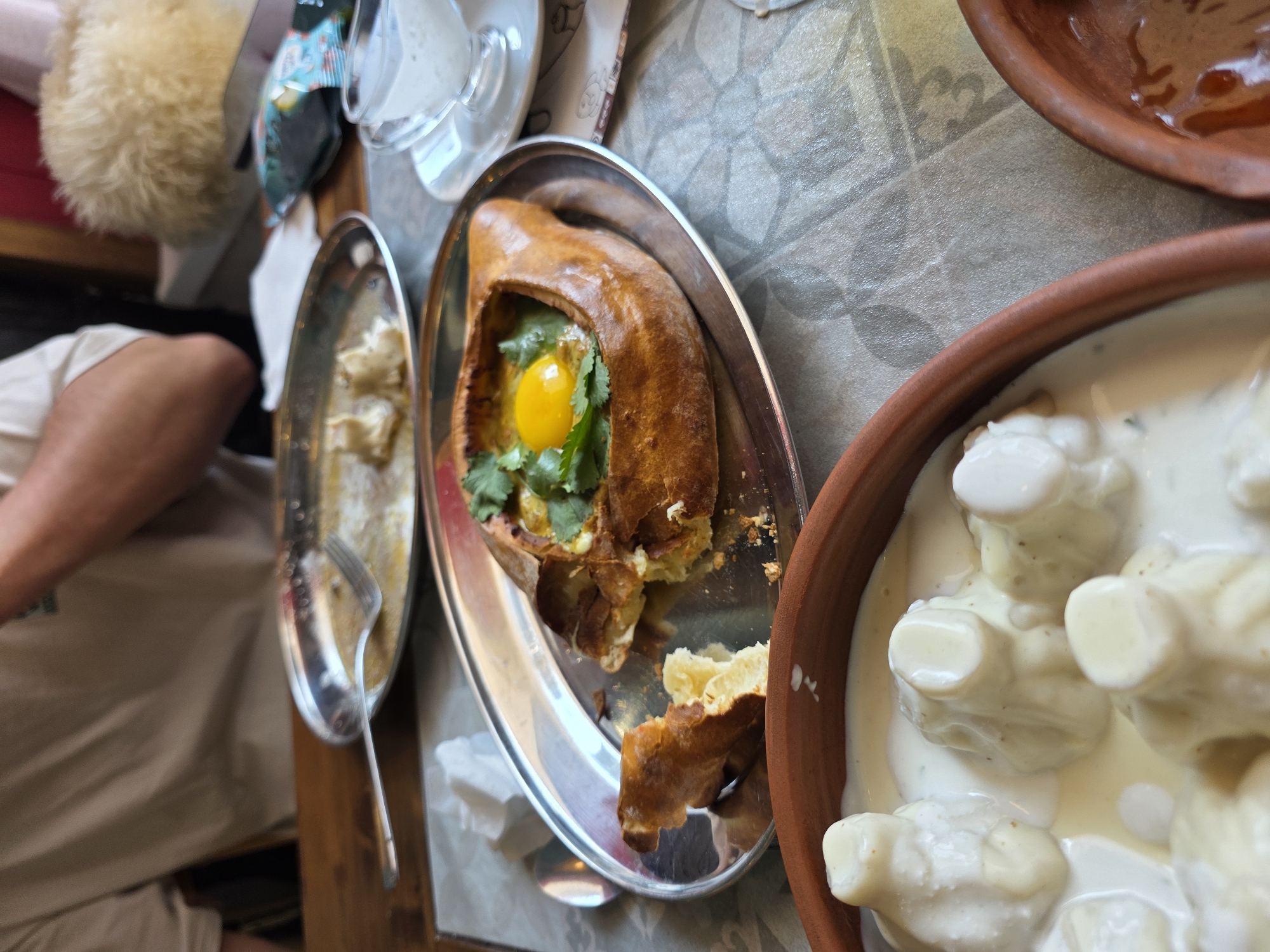 A high-angle, close-up shot captures a table setting in a restaurant, likely in Sochi, Russia, with two dishes of Georgian food prominently displayed. In the foreground, a terracotta bowl contains several dumplings swimming in a creamy white sauce. To the right and slightly behind, a silver oval plate holds a Khachapuri Adjaruli, a boat-shaped bread filled with cheese and topped with a vibrant yellow egg yolk and fresh green herbs. A piece of the bread's crust is broken off and rests on the plate. A small glass gravy boat filled with a white sauce sits to the right of the Khachapuri. In the background, a person wearing a beige t-shirt is partially visible, with their hands resting on the table. The table is covered with a textured grey tablecloth. Additional elements like a fork on an empty silver plate, a crumpled napkin, and a packet of what appears to be food seasoning are also present, suggesting a meal in progress. The overall atmosphere is casual and inviting, indicative of a dining experience.