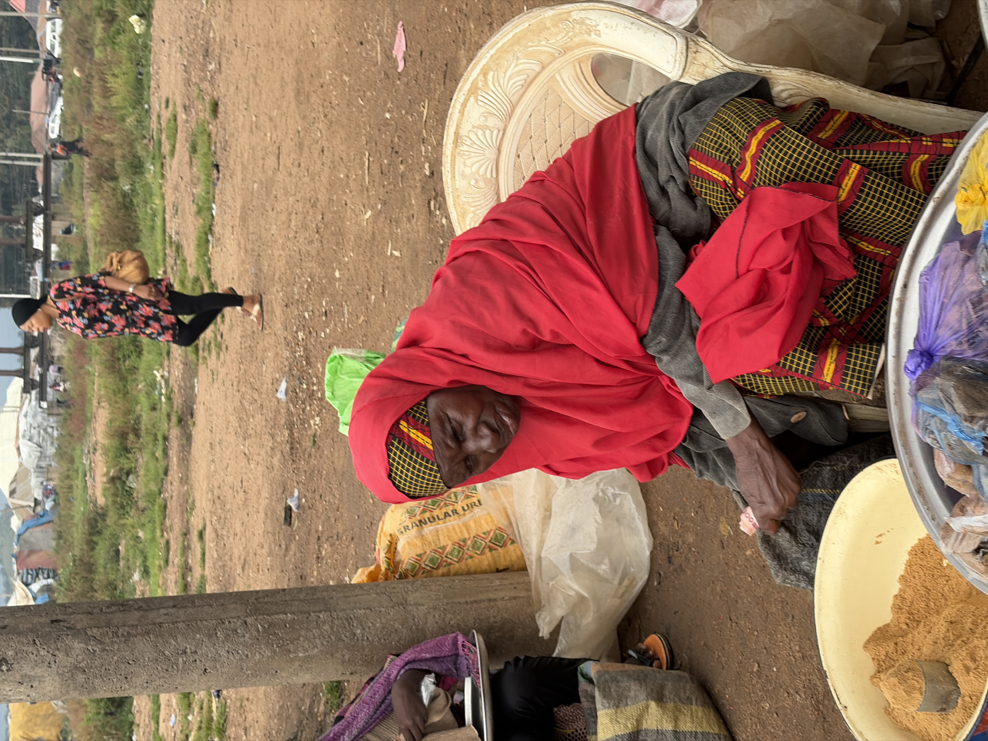 The image depicts an outdoor market scene in Bukuru, Nigeria. In the foreground, an elderly woman wearing a vibrant red headscarf and a patterned skirt is seated. Her expression appears weary as she holds a small pink object, possibly currency, in her hand. To her left, another person, partially visible, is seated near a pile of goods. A large yellow bag with red and green diamond patterns is visible behind the seated woman. The ground is a mix of dirt and sparse vegetation. In the background, tents and temporary structures suggest a makeshift settlement or a large market area. A woman in a floral dress and black leggings walks in the midground, carrying a brown bag over her shoulder. Other individuals and vehicles are faintly visible in the distance, indicating a bustling environment. The overall atmosphere suggests a busy, informal trading post within the city.