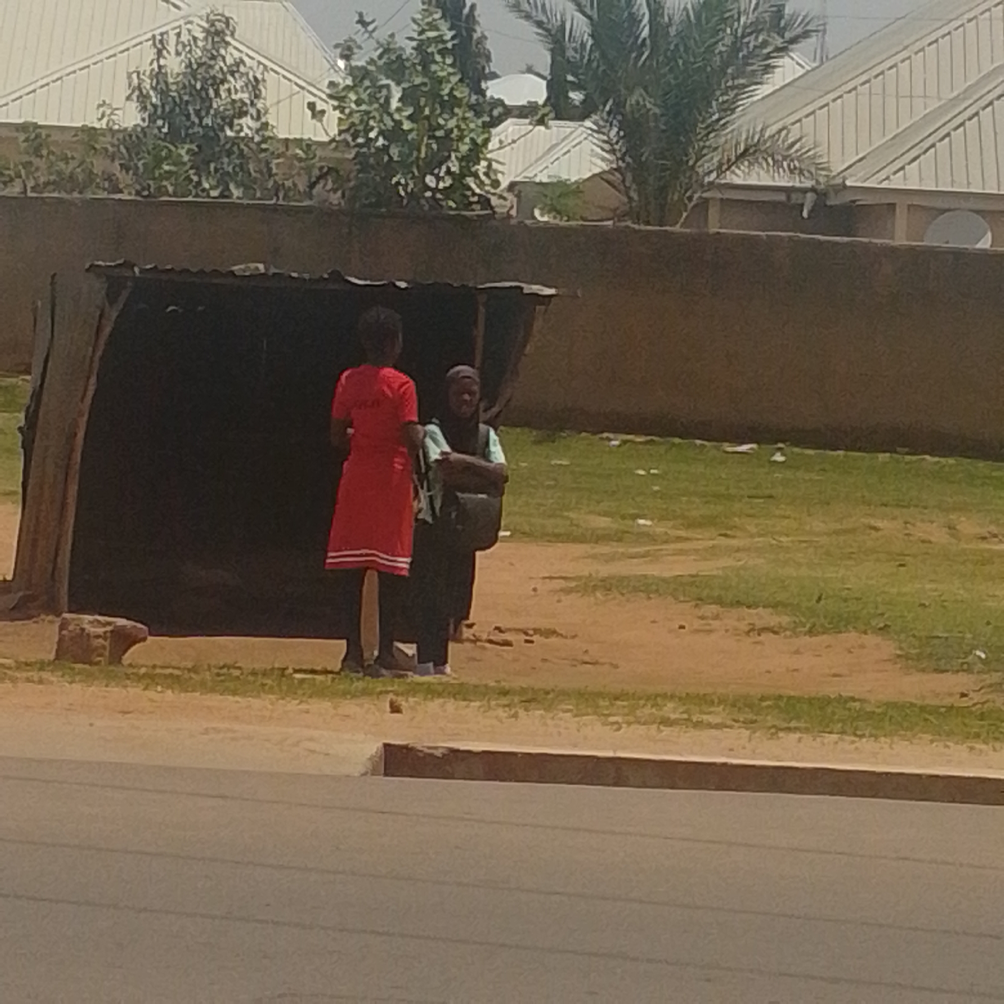 Two individuals are standing near a makeshift shelter by a road. The shelter appears to be constructed from corrugated metal and possibly some wooden poles or branches. On the left, a person is wearing a red dress with white stripes at the hem and standing with their back to the camera. To their right, a second person is wearing a dark head covering and a light-colored top, with arms crossed. They are both standing on a dirt path next to the shelter. Behind the shelter, there is a grassy area and beyond that, buildings with white, sloped roofs, and some trees. The scene is set in Tempure, Nigeria, a location characterized by its arid climate and residential areas with distinct architectural styles, evident in the white-roofed structures and palm trees visible in the background.
