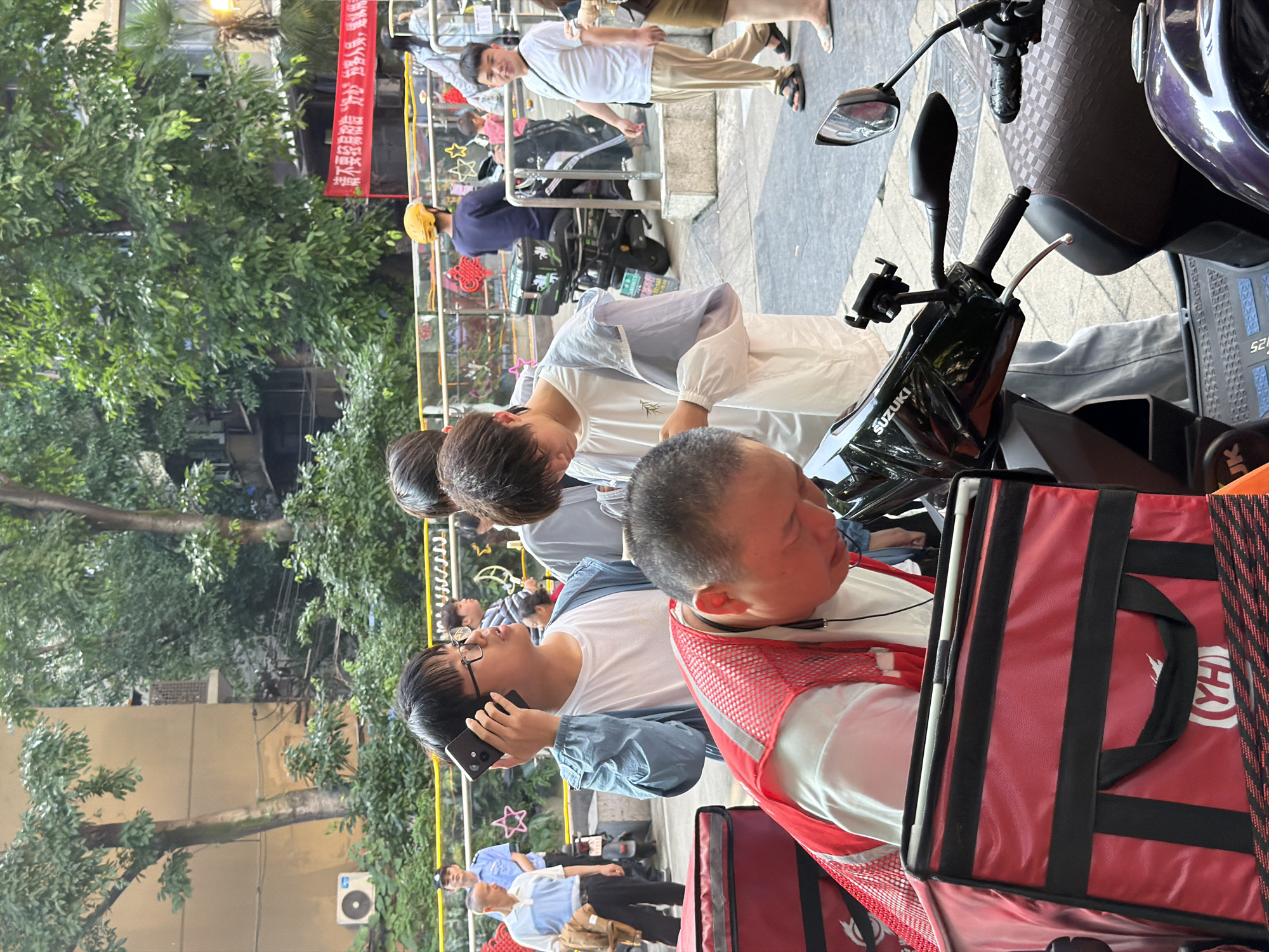 In the Yuzhong District of China, an outdoor scene unfolds with a focus on delivery personnel and individuals in an urban environment. In the foreground, a man wearing a red mesh vest and a white shirt is leaning over, tending to a large red delivery bag. To his left, another red delivery bag is visible. A black Suzuki scooter is positioned prominently, with its handlebars and a phone mount in view. Behind this, a younger man in a light blue jacket and white t-shirt is holding a black smartphone to his ear, appearing to be on a call. Next to him, another individual in a light colored jacket is looking down at something. In the middle ground, a person wearing a yellow helmet is seated on a scooter. Further back, a man in a light blue police uniform stands near an older man wearing a white polo shirt and a brown jacket. A red banner with Chinese characters is stretched across the background. Green trees and buildings with visible air conditioning units form the backdrop. The ground is paved with grey tiles.
