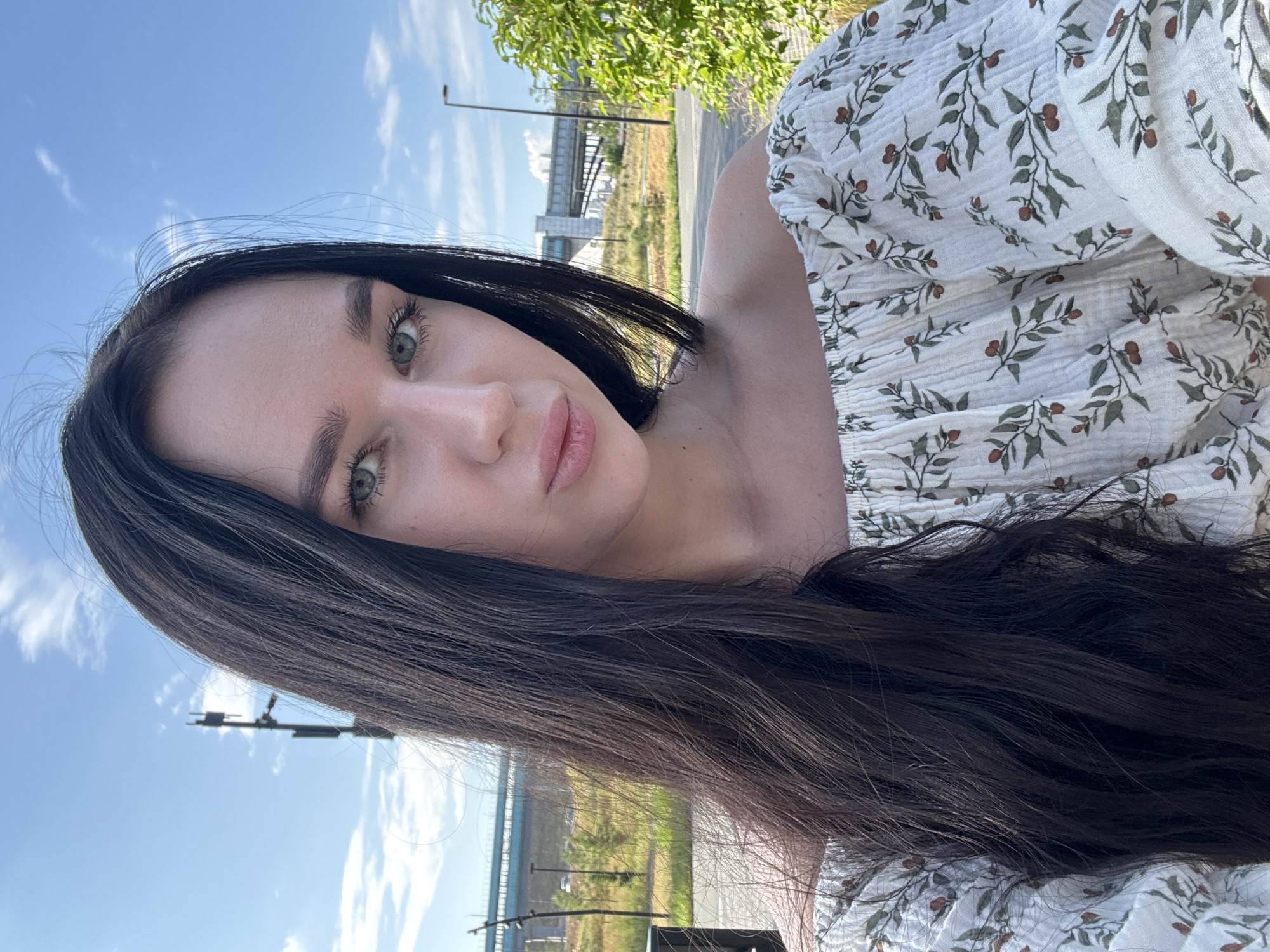 A young woman with long, dark hair is taking a selfie outdoors on a sunny day. She is wearing an off-the-shoulder white blouse with a floral print. She is looking at the camera with a neutral expression. In the background, there is a blue sky with white clouds, a cell tower, a bridge, trees, and a grassy field. The setting appears to be a park or an open area on the outskirts of the city. The image does not provide specific details to pinpoint the exact location within Novosibirsk, Russia.