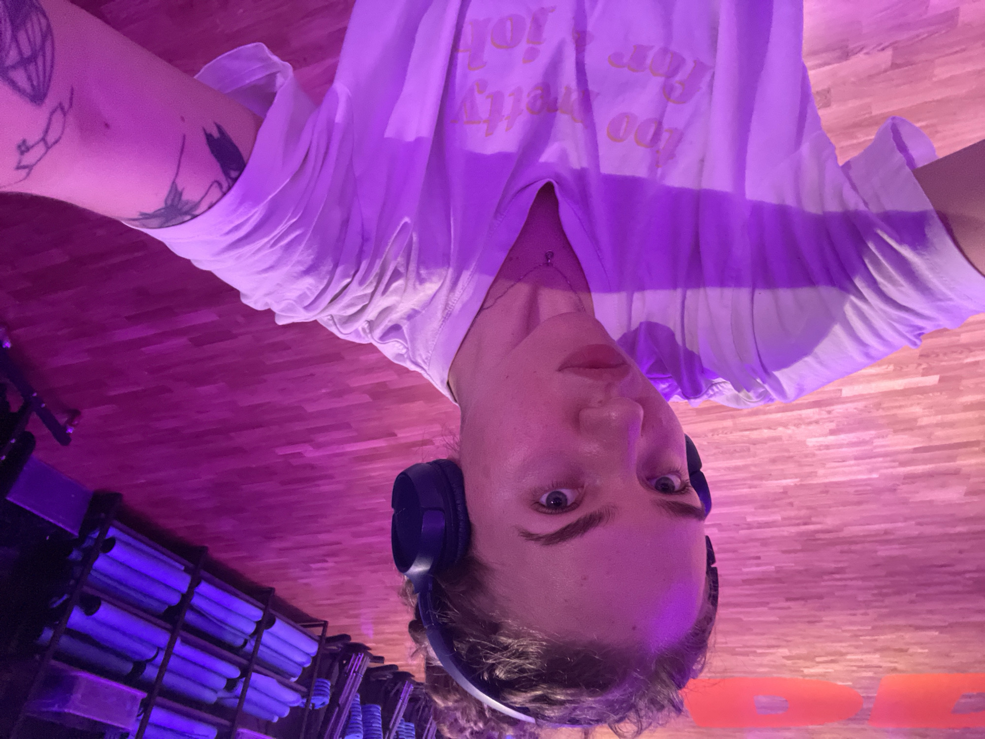 A person is looking down at the camera, wearing headphones and a white t-shirt with lettering on it. The environment is lit with purple light, and there is a wooden floor. A rack of rolled-up mats is visible in the background. There is no indication that this image was taken in Moscow, Russia.