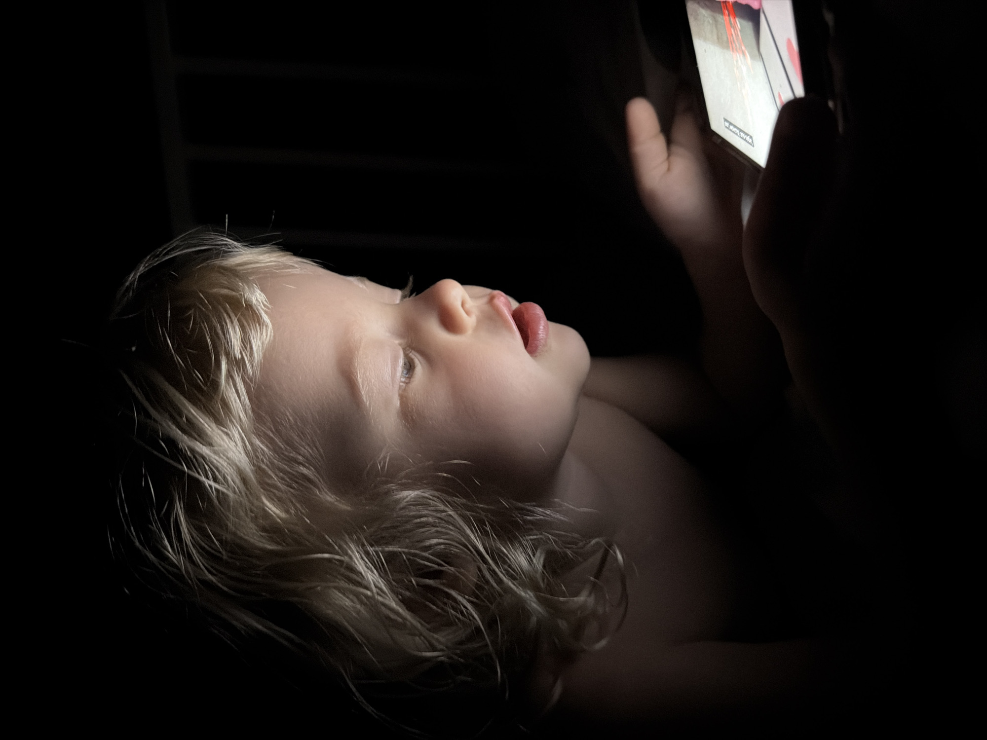 A young child, with light, wavy hair and bare shoulders, is observed in a dark room in the city of Sochi, Russia. The child's face and hands are brightly illuminated by the screen of a smartphone or tablet device, which the child holds and intently views. The screen displays active content with prominent red and white visual elements. The child's mouth is slightly ajar, indicating concentration. The surrounding environment is almost entirely dark, with minimal ambient light, though a faint structural element, possibly a bed rail, is visible in the upper right background.