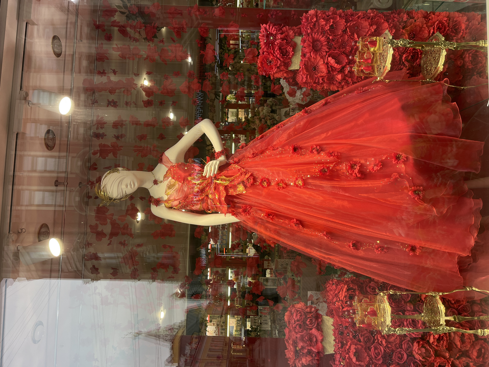 A red dress on a mannequin is displayed in a store window in Moscow, Russia. The dress is surrounded by red flowers. The window reflects the surrounding city.