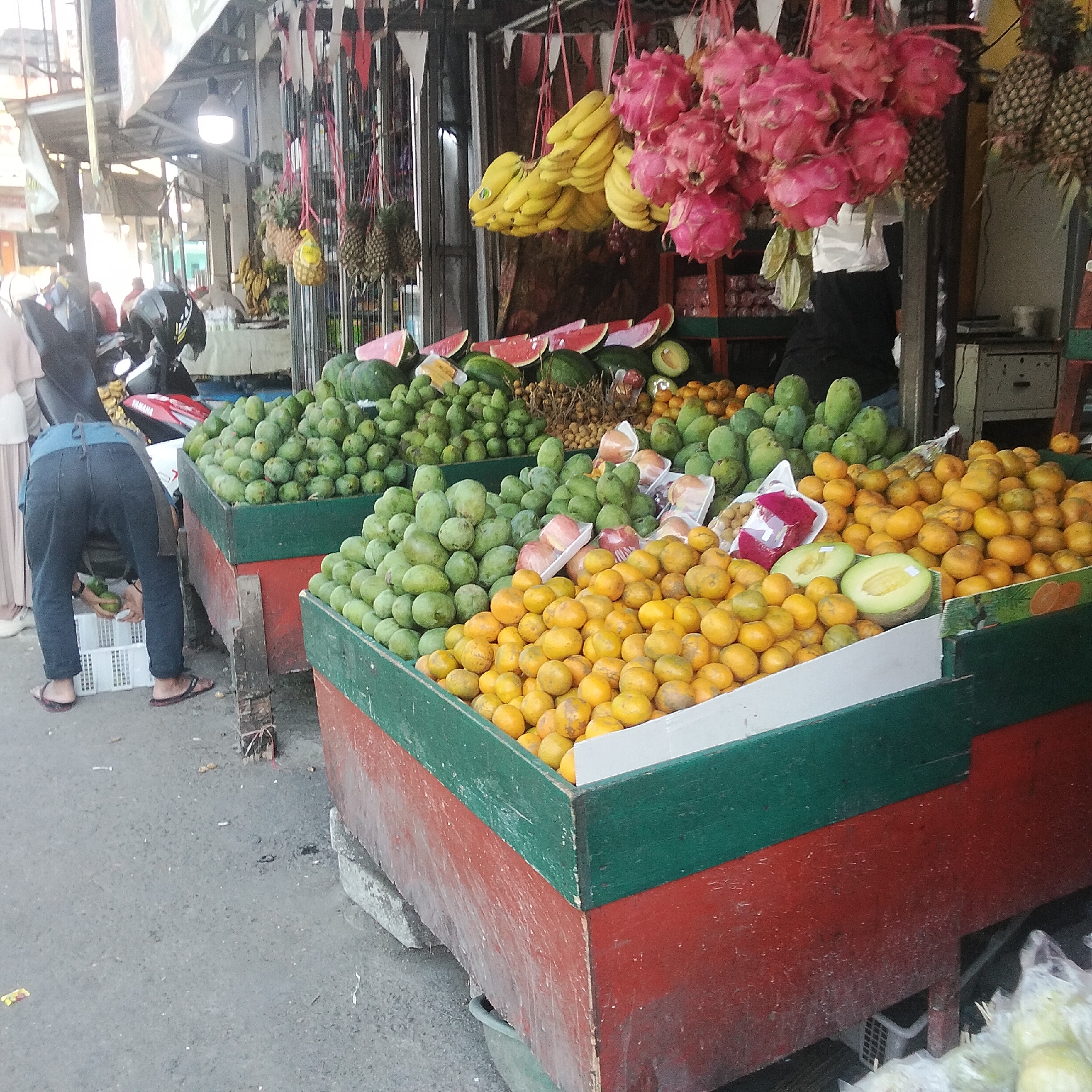 This is a medium shot of a fruit stall in Sukasari, Indonesia. The stall is made of wood and painted red and green. In the foreground, there are two large bins filled with fruit. The bin on the left is filled with green mangoes. The bin on the right is filled with oranges. Behind these bins, there are more fruits visible including watermelons, dragon fruits, pineapples, and bananas. In the background, a person wearing a blue shirt and dark pants is bending over and appears to be handling some of the fruits. Another person wearing a light-colored headscarf and a light-colored dress is standing near a motorcycle. The scene appears to be outdoors, on a paved surface.