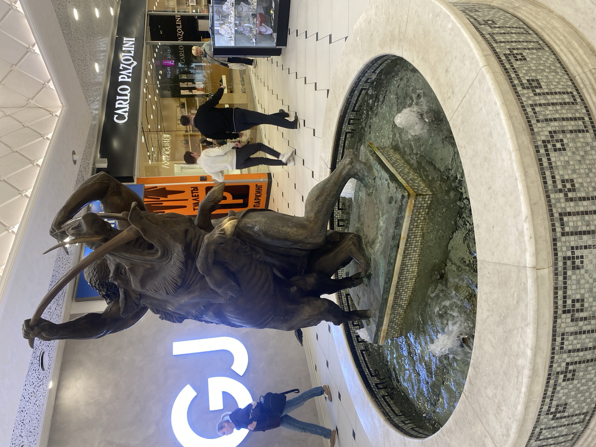 Here's a detailed description of the image:
**Location and Environment:**
The image appears to be taken inside a shopping mall or commercial center in Yekaterinburg, Russia. A prominent feature is a large, circular fountain. The fountain has a decorative mosaic rim and contains a bronze sculpture.
**Subjects and Objects:**
* **Bronze Sculpture:** A bronze sculpture of a man fighting a large animal (possibly a boar or bull) is positioned within the fountain.
* **Fountain:** The fountain has running water and is surrounded by white and mosaic tiles.
* **People:** Several people are present in the scene. One woman stands closer to the fountain, looking in the general direction of the sculpture. Other people appear to be walking past shops in the background.
* **Shops:** The background shows shop fronts. One visible sign reads "CARLO PAZOLINI", a brand store. Another sign with an orange background has text in Russian, including the word "ПАРКИНГ" (Parking).
* **Signage:** A large, illuminated "GU" sign is visible near the woman.
* **Flooring:** The mall floor is tiled.
**Actions and Interactions:**
* People are walking through the mall, seemingly shopping or passing by.
* The woman standing near the fountain may be looking at or admiring the sculpture.
* Water is flowing in the fountain.
**Overall Impression:**
The image captures a moment in a shopping center, where people interact with the commercial environment and a central artistic feature—the fountain and sculpture.