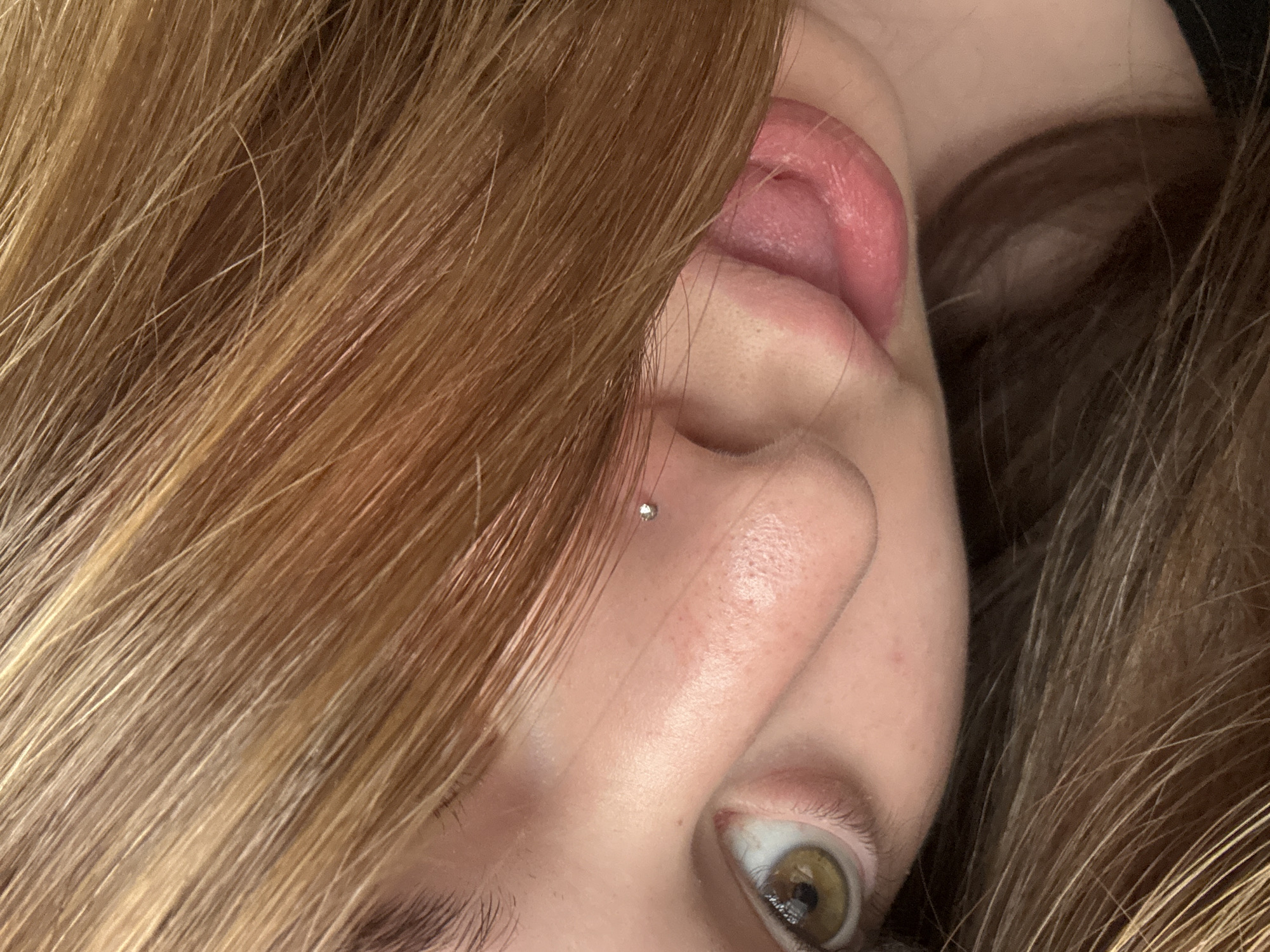 This close-up, vertically oriented photograph captures a segment of a person's face, rotated approximately 90 degrees to the left. The subject's light brown or reddish-blonde hair dominates the left and upper portions of the frame, appearing textured and slightly wavy.
The right side of the face is visible, including a prominent right ear with a pinkish inner canal, suggesting the earlobe is angled upwards. A small, shiny silver stud piercing is visible on the bridge of the nose, positioned relatively high between the eyes. Below the piercing, the person's right eye is open, revealing a light brown or hazel iris with a dark pupil, reflecting a point of light. The eyebrow is partially visible at the bottom of the frame.
In the upper right corner, a glimpse of pinkish flesh, possibly the upper lip, inner cheek, or tongue, is visible near the ear, suggesting the mouth might be open or in motion. The skin tone is fair with some slight texture visible. The image appears to be a tightly cropped portrait, possibly a selfie, focusing on the facial features and the piercing. No specific background details, time of day, weather, or visible text can be discerned from the image. The general impression is one of a personal and intimate shot.