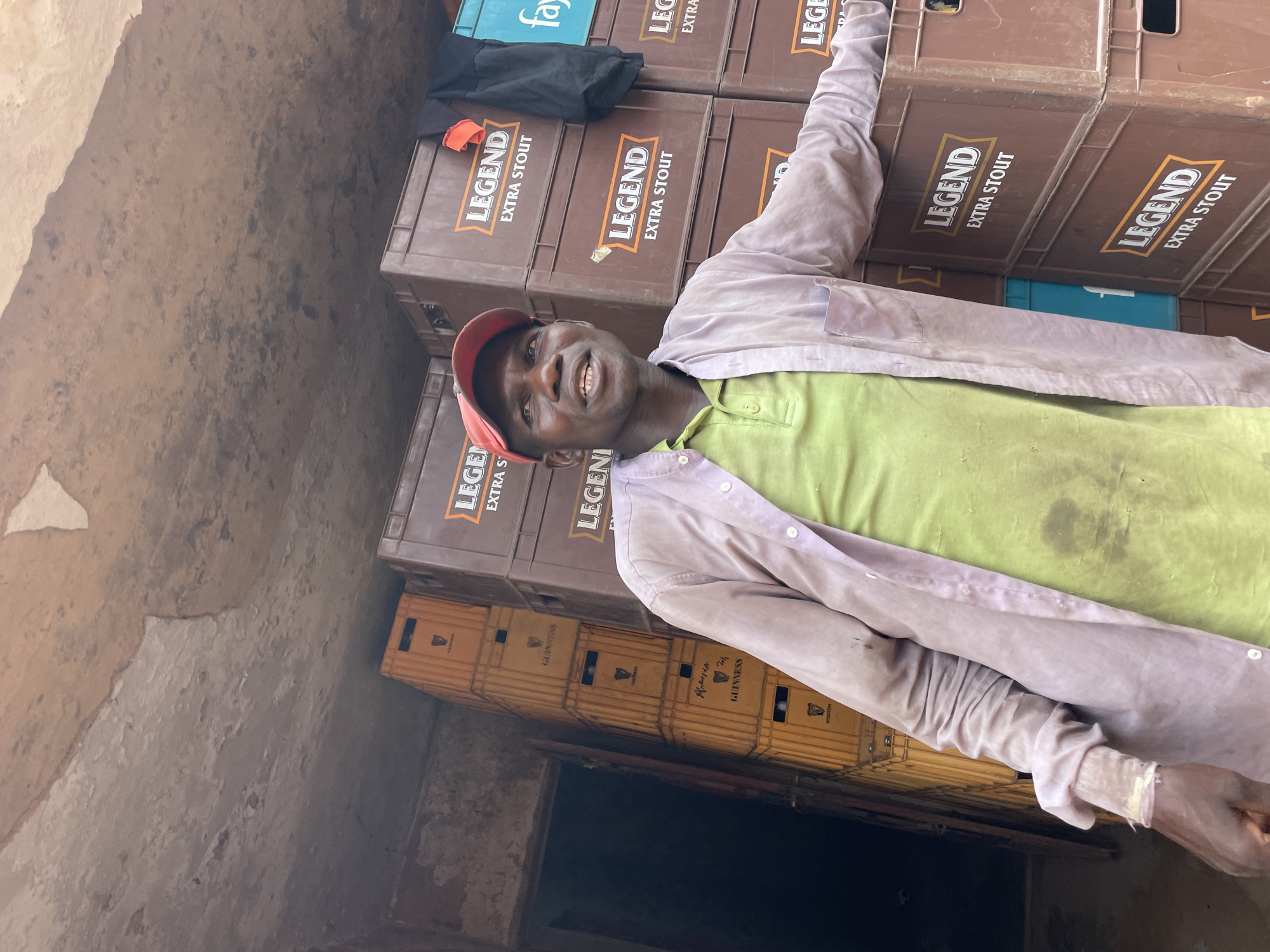 The image depicts a man leaning against a stack of beer crates in what appears to be a storage area or warehouse. The crates are primarily for "Legend Extra Stout" and some crates for "Guinness", stacked in neat rows. The man, who is smiling, wears a red cap, a light green shirt, and a light-colored long-sleeved shirt or jacket. The lighting in the area is somewhat dim. The walls of the storage area are rough and uneven. The image was taken in Bukuru, Nigeria.