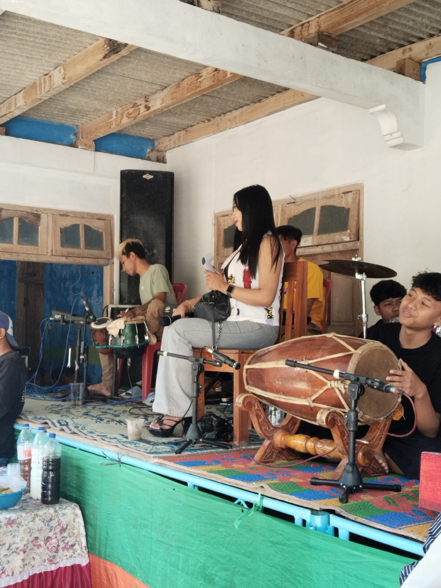 A medium shot shows a performance with musicians and a singer on an elevated stage.
A woman with long black hair is seated in a wooden chair, holding a microphone. She wears a white top with a graphic design and light grey pants. She is slightly turned, facing towards other members of the band.
To the left, a man with dyed blond hair sits behind a pair of percussion instruments. He wears a light green shirt. Behind the woman, another man in a yellow shirt is partially visible. To the right, a young man in a black shirt is seated and playing a traditional Indonesian drum with a microphone in front of it. A cymbal on a stand can be seen slightly behind them.
Behind the musicians are windows with wood framing and a black speaker.
The stage has a blue frame, with a green and orange fabric covering the front. A table covered with a floral tablecloth is on the left side, holding bottles of water and other items. The floor of the stage is covered with a patterned rug.
The location appears to be indoors, under a corrugated iron roof supported by wooden beams. The walls are painted white.